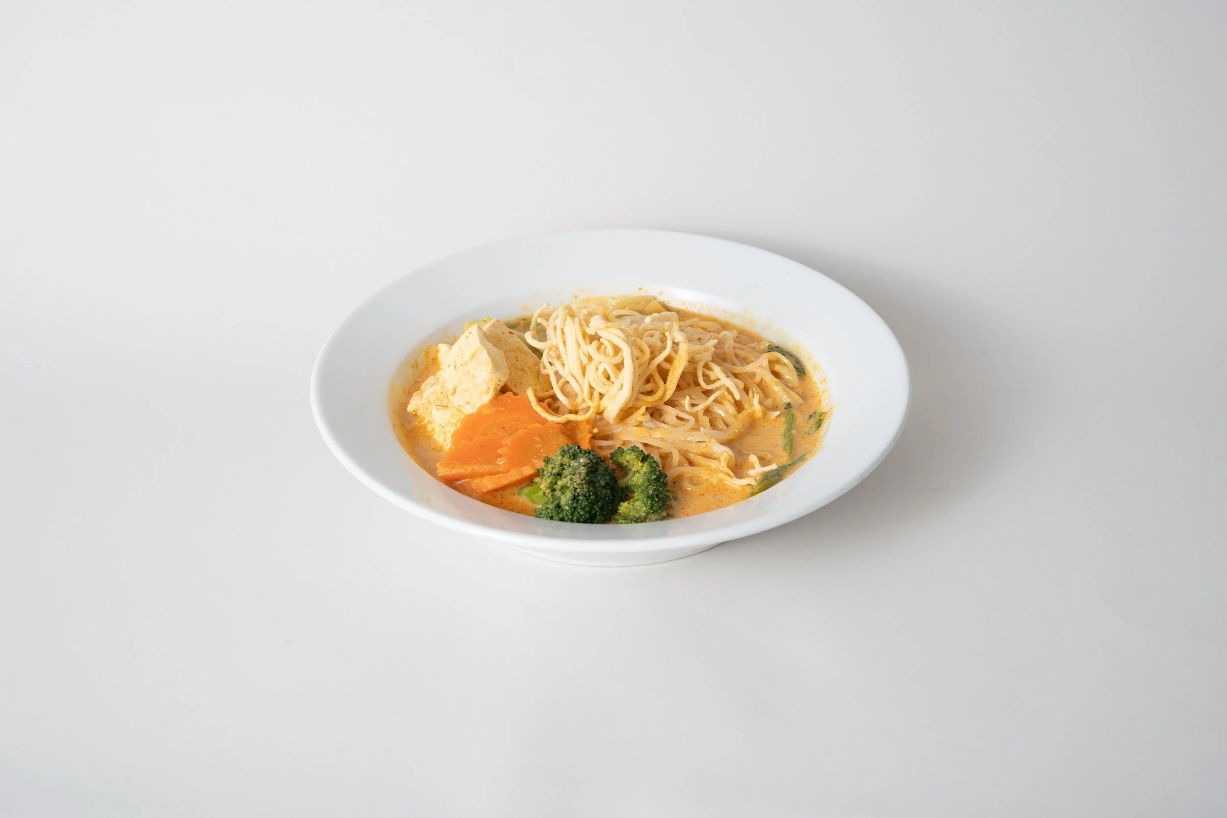 A white bowl of ramen noodles with vegetables including broccoli, carrots, and tofu in a creamy broth on a white surface.