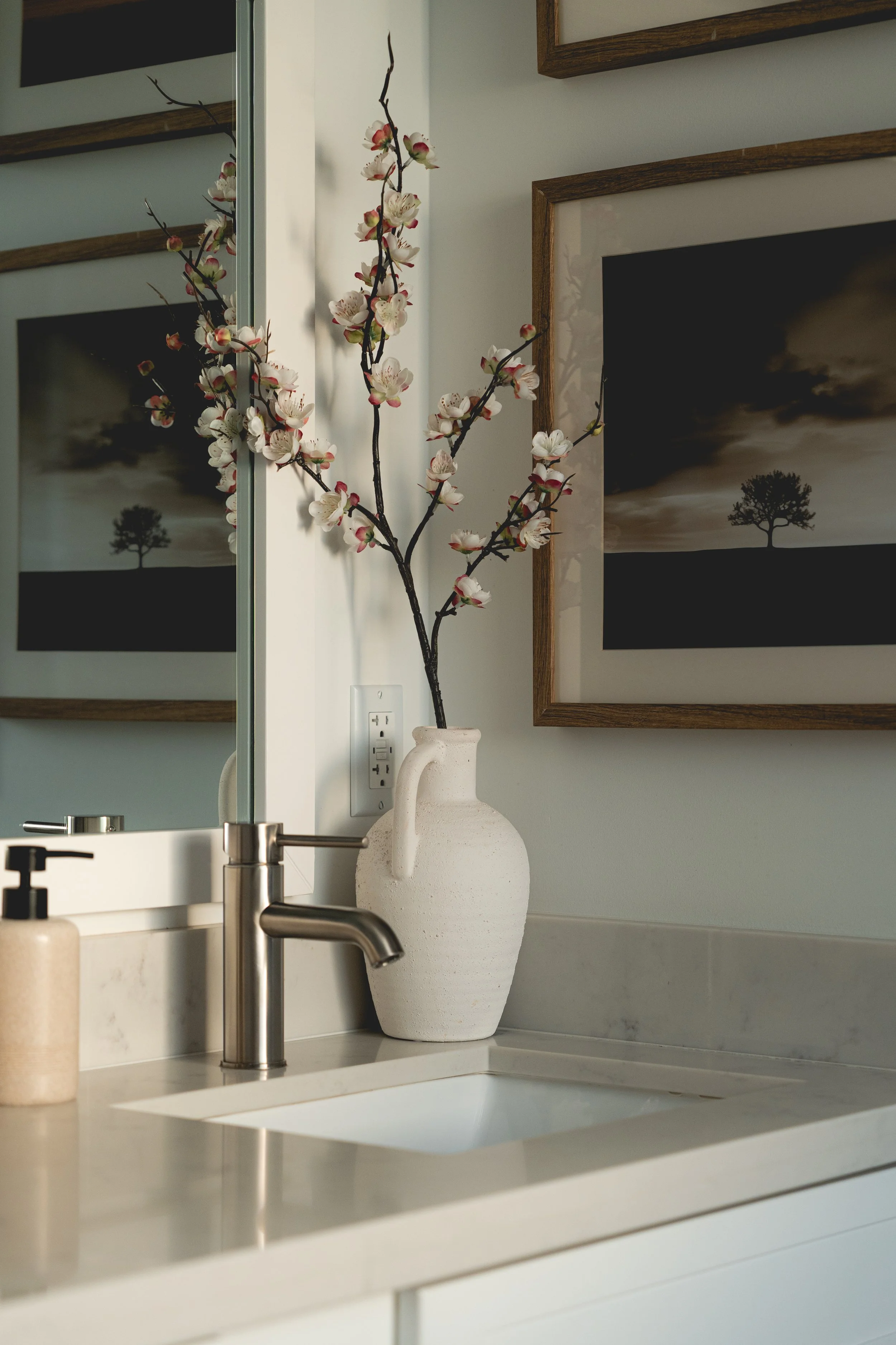 Bathroom countertop with sink, a vase with pink and white blossoms, a mirror, and framed artwork of a tree silhouette.
