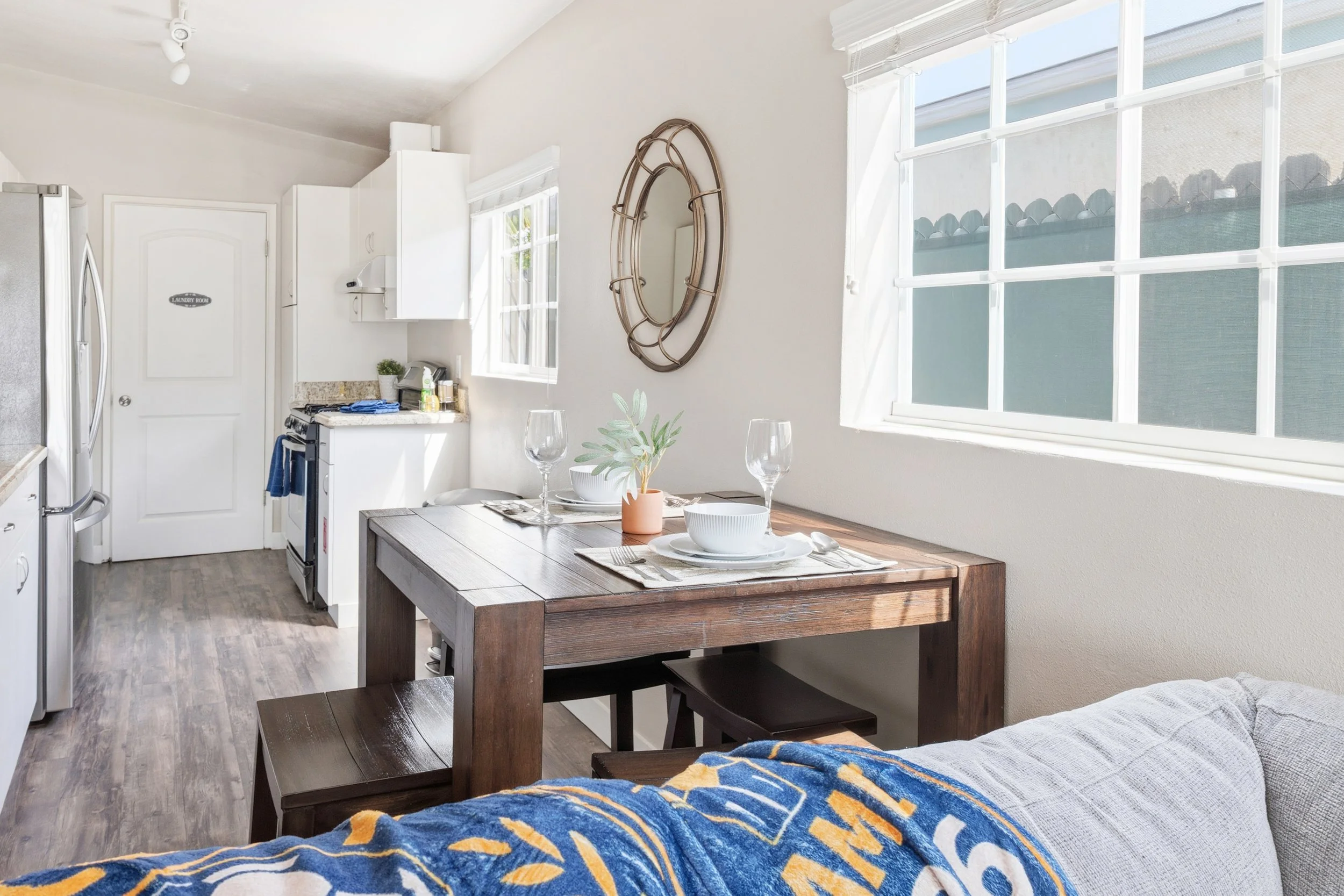 A cozy kitchen and dining area with a wooden table set for two, including plates, bowls, wine glasses, and a small plant. The room has white walls, large windows with blinds, and a small wall mirror. In the background, there is a white door with a si