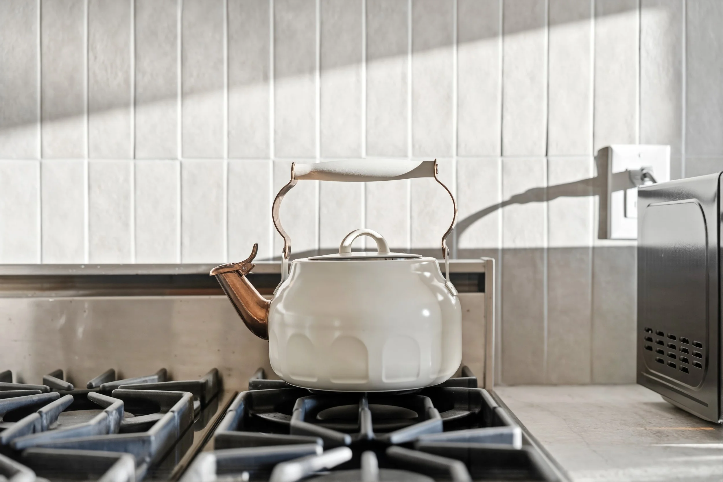 A white kettle with a copper handle on a stovetop in a kitchen with white tiled walls and a gray countertop.