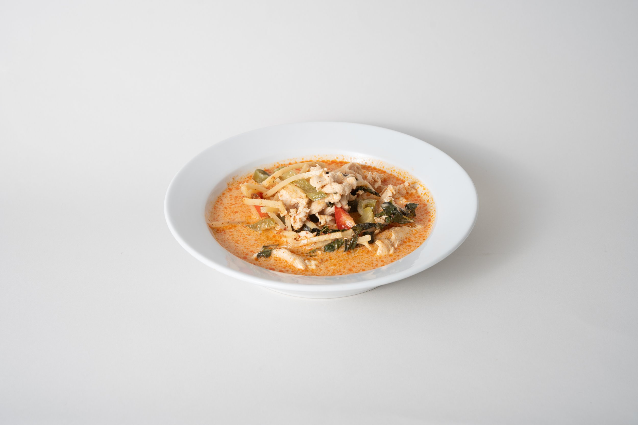 A white bowl of Thai spicy chicken soup with vegetables and herbs on a white surface.
