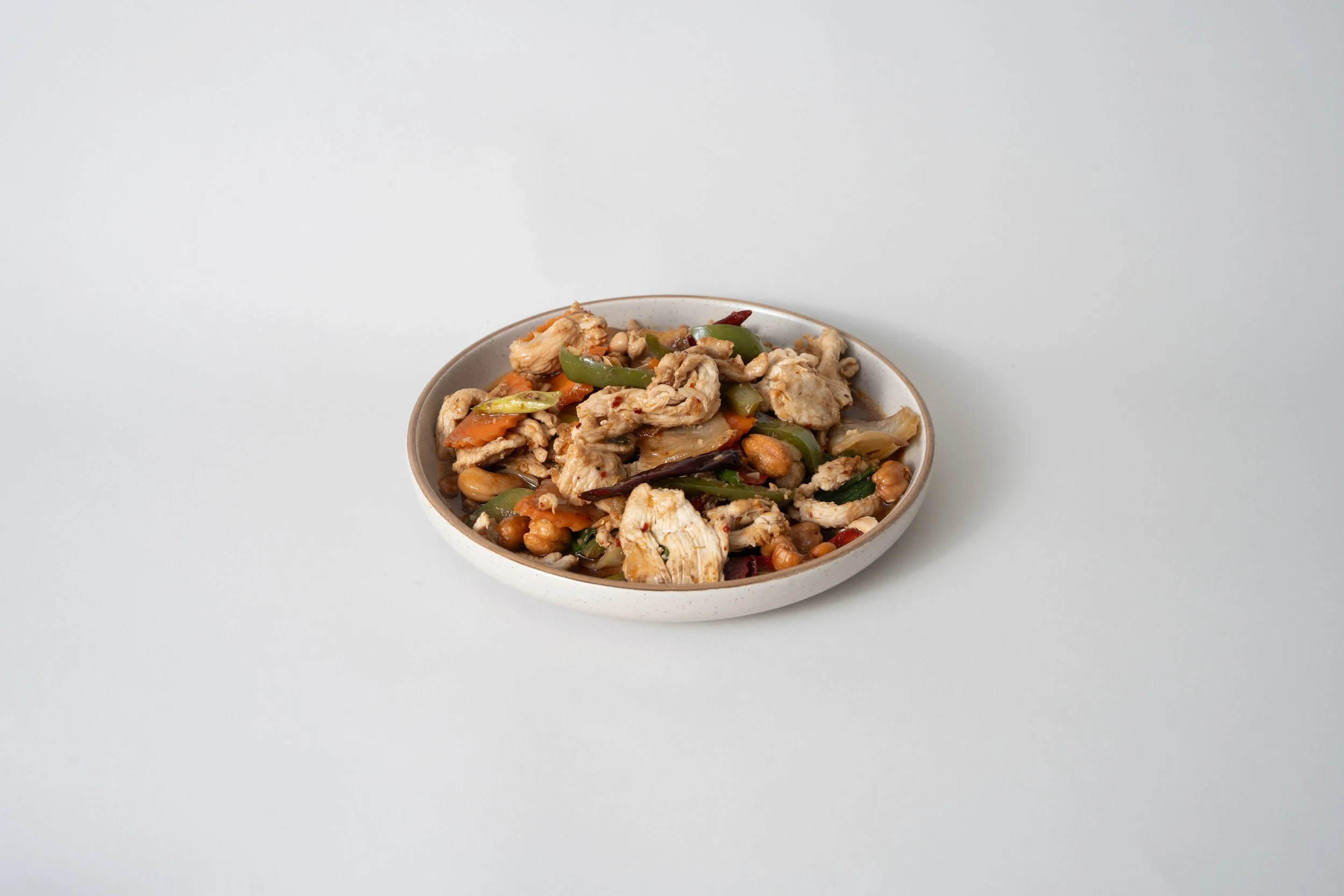 A bowl of stir-fried chicken with vegetables and peanuts on a plain white background.