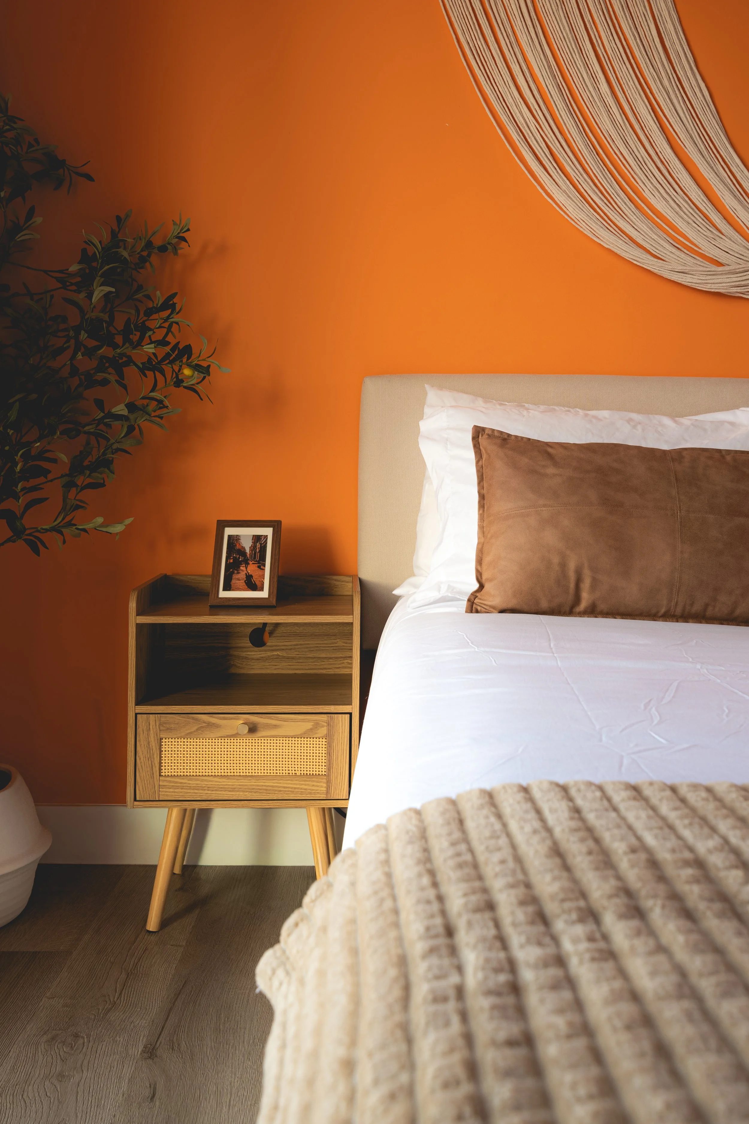 A cozy bedroom with an orange accent wall, a beige headboard, white linens with a brown pillow, a textured beige throw blanket, a wooden nightstand with a small framed picture, a potted plant, and a large woven wall hanging.