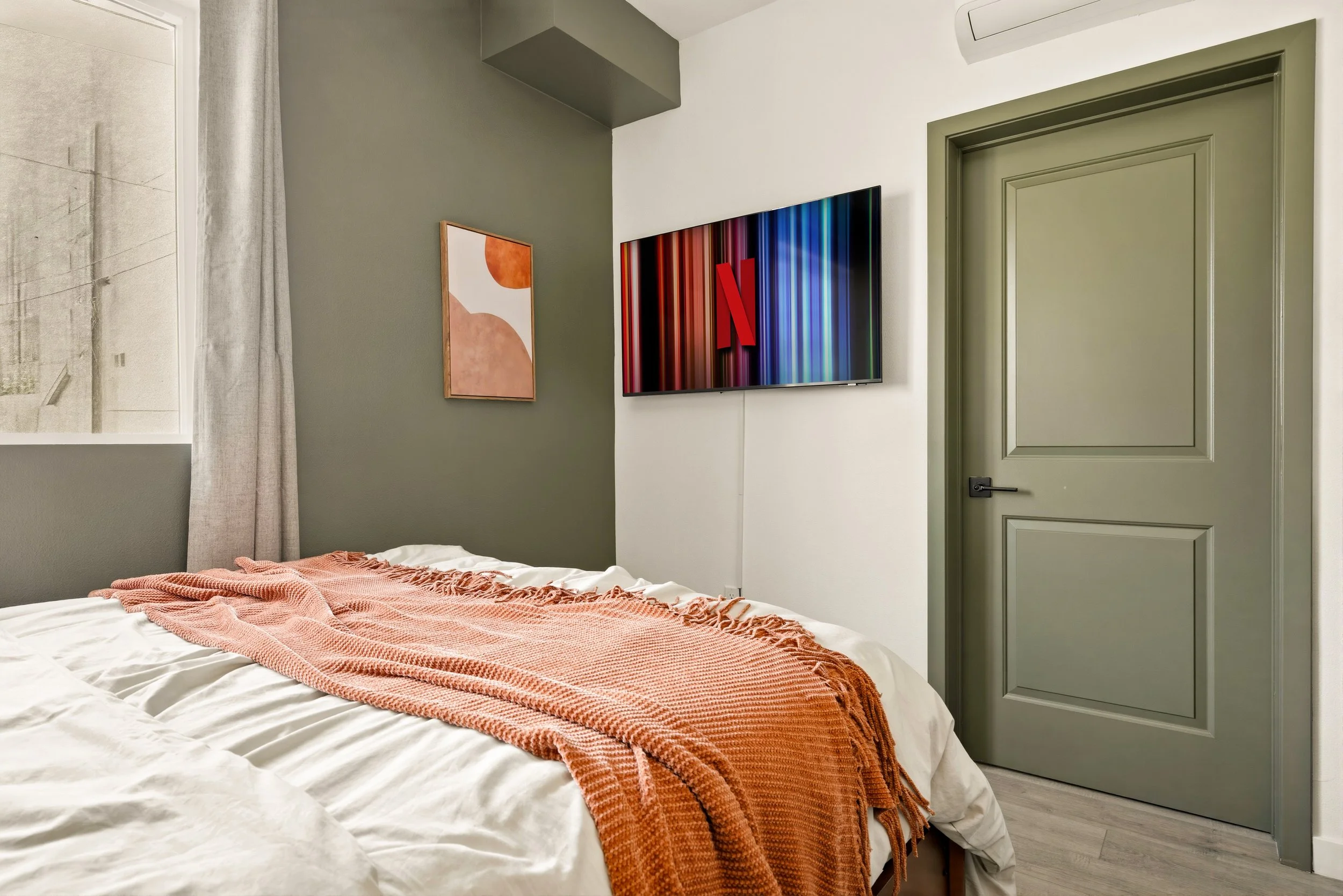 A bedroom with a bed covered in white sheets and a peach-colored throw blanket, a green door, a window with a curtain, framed artwork on the wall, and a TV on the wall displaying the Netflix logo.