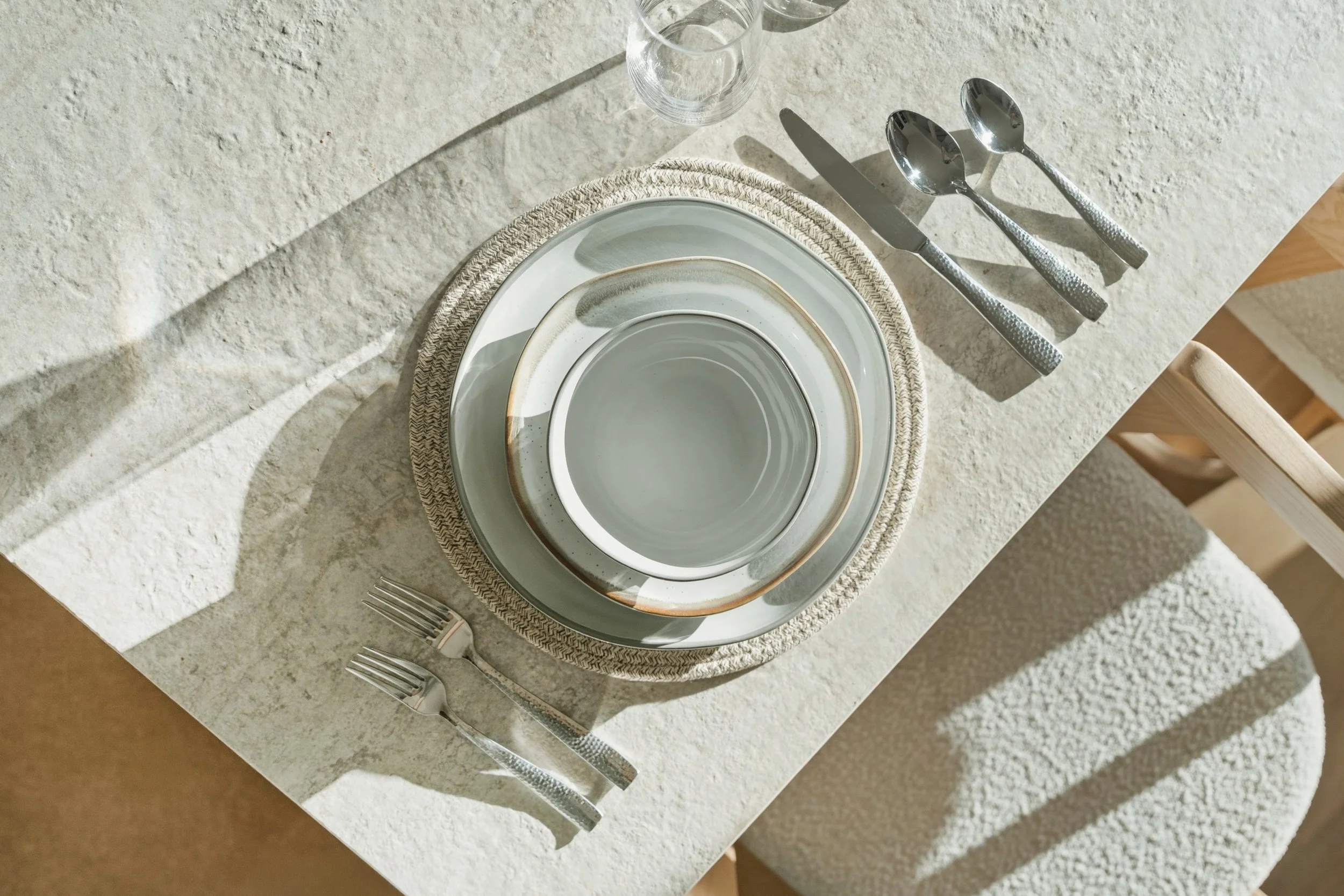 Stacked ceramic plates on a woven placemat set on a white textured table, with silver cutlery and two empty glasses nearby, under natural sunlight.