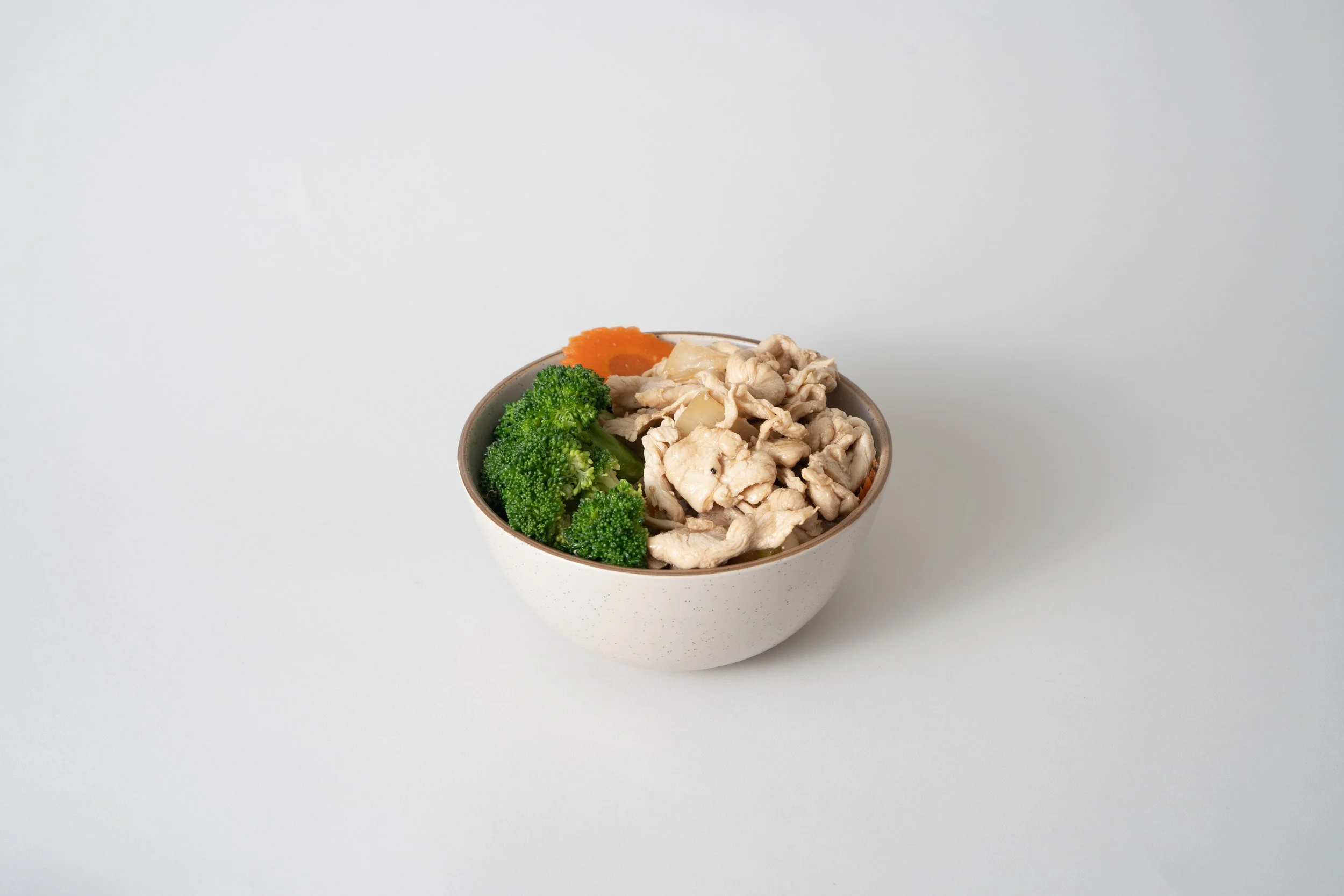 Bowl of cooked broccoli, shredded chicken, and a slice of carrot on a white background.