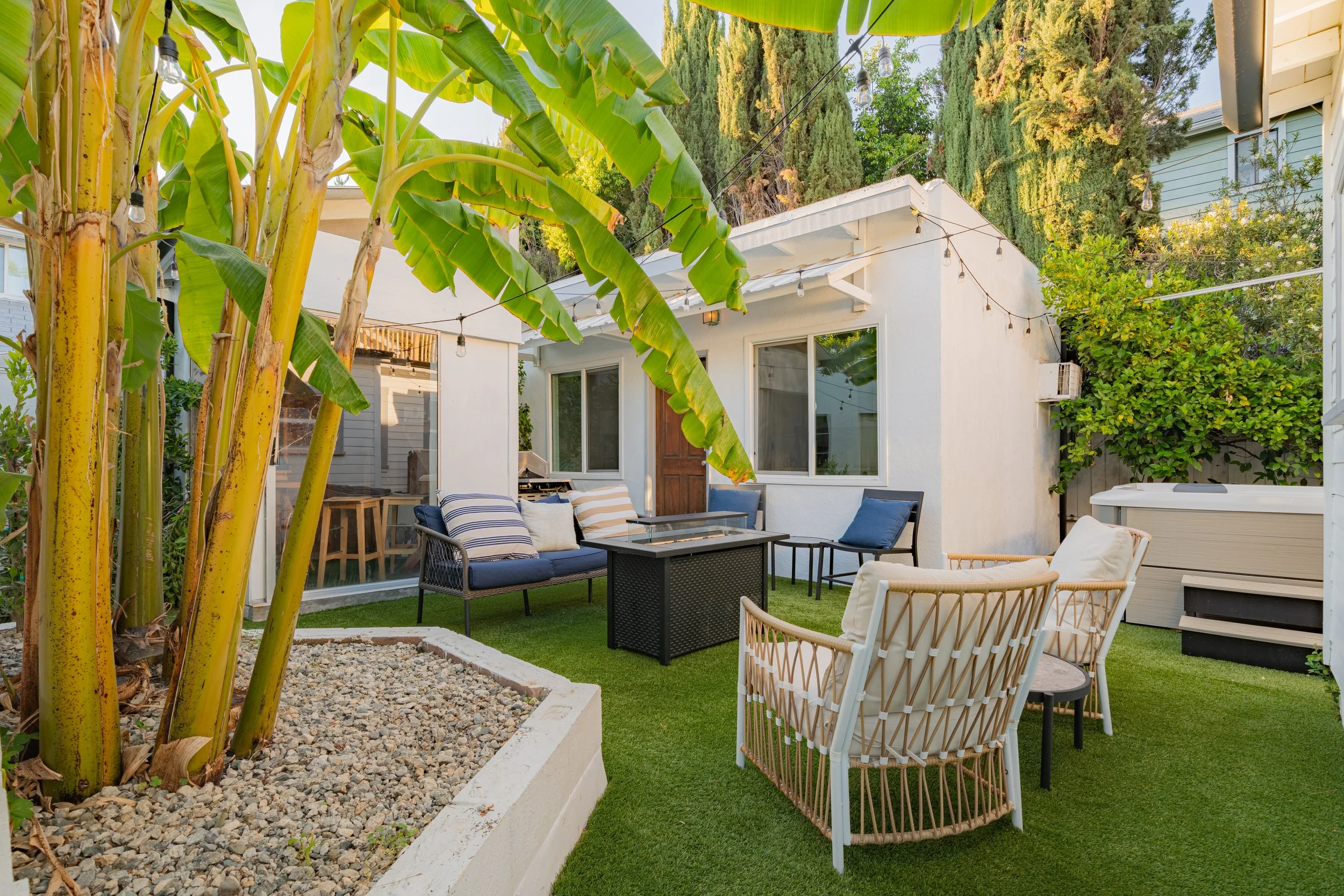 A cozy outdoor backyard patio with seating and string lights, featuring banana plants in a landscaped garden with a white house in the background.