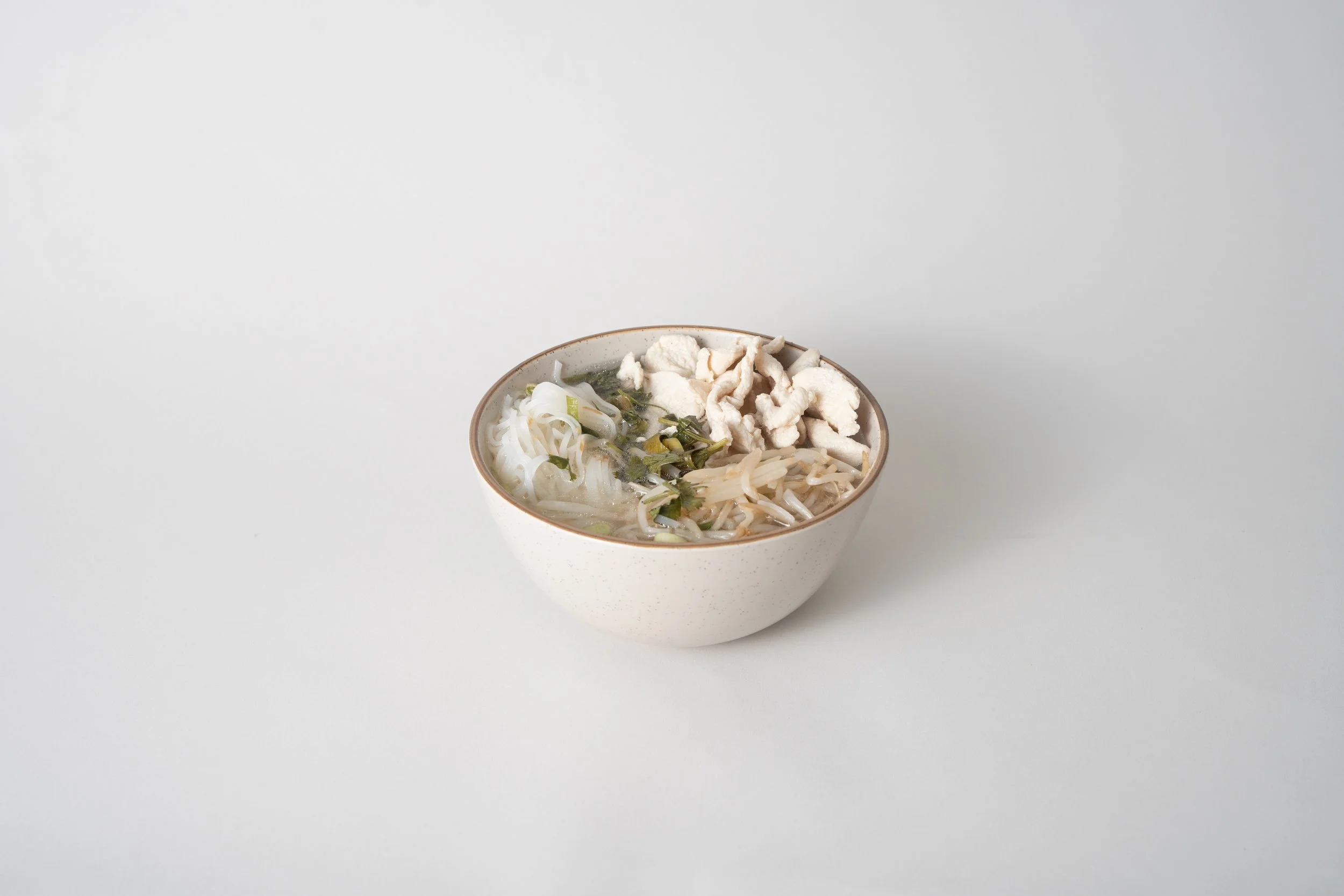 A white bowl with Asian-style noodle soup topped with white shredded chicken, herbs, and vegetables on a plain white background.