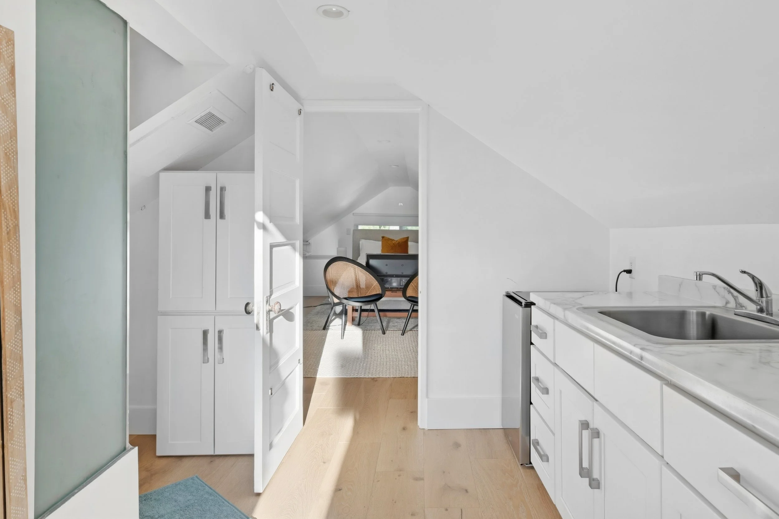 View of a white kitchen with a sink, countertop, cabinets, and a doorway leading to a dining area with chairs and a bed visible in the background.