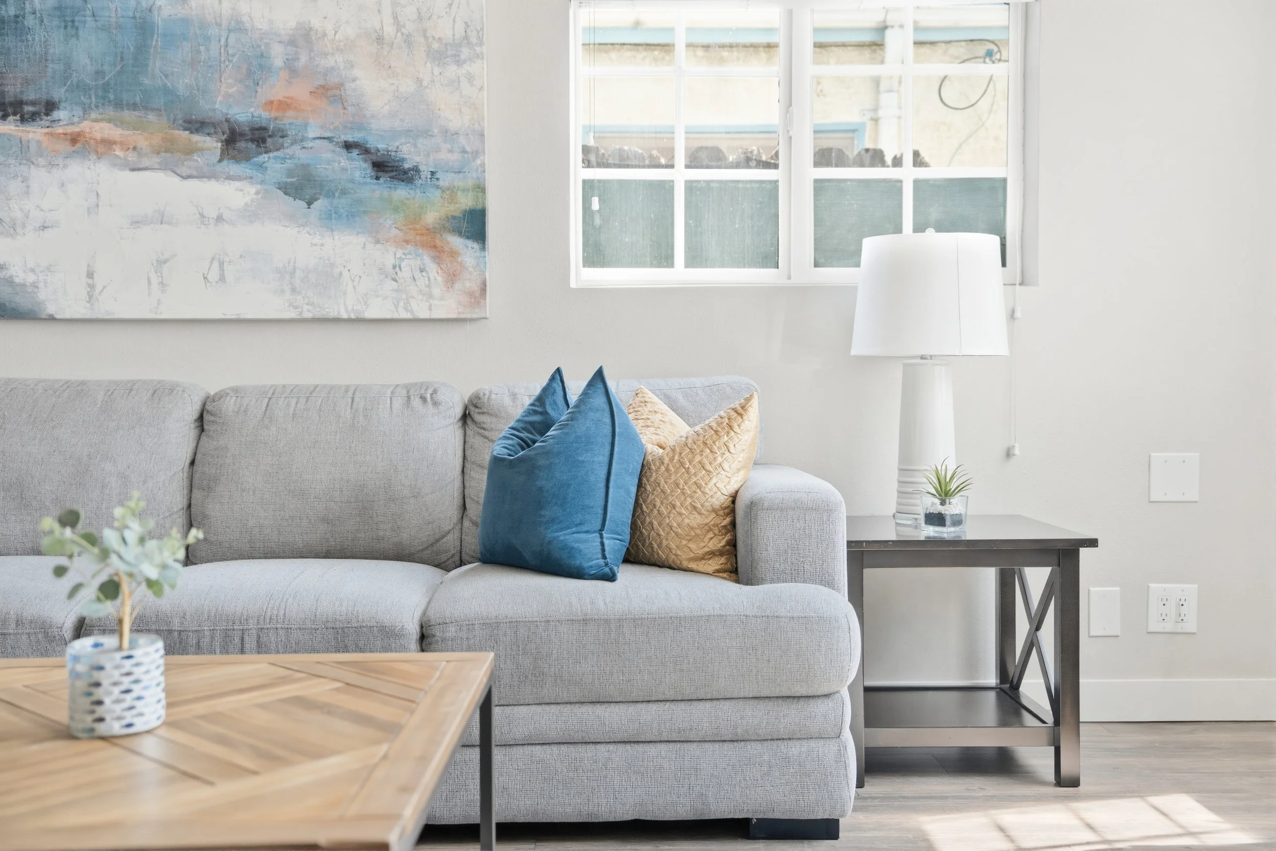 Living room with light gray sofa, blue and beige throw pillows, wooden coffee table, black side table with a small plant and white lamp, abstract art on the wall, window with sunlight streaming in.