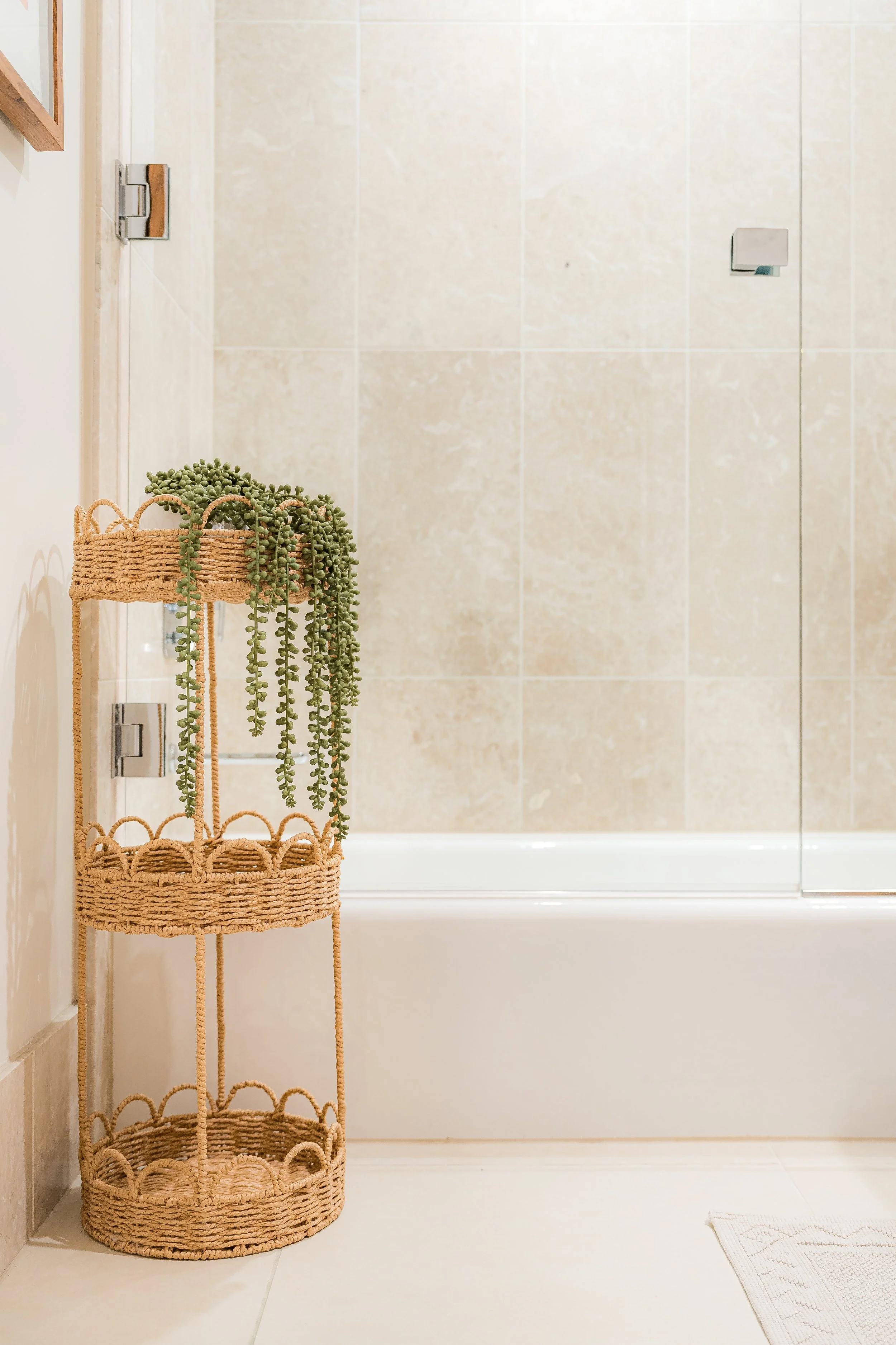 A wicker plant stand with potted succulent on top, situated in a bathroom with a bathtub, beige tiled wall, and a small towel on the floor.