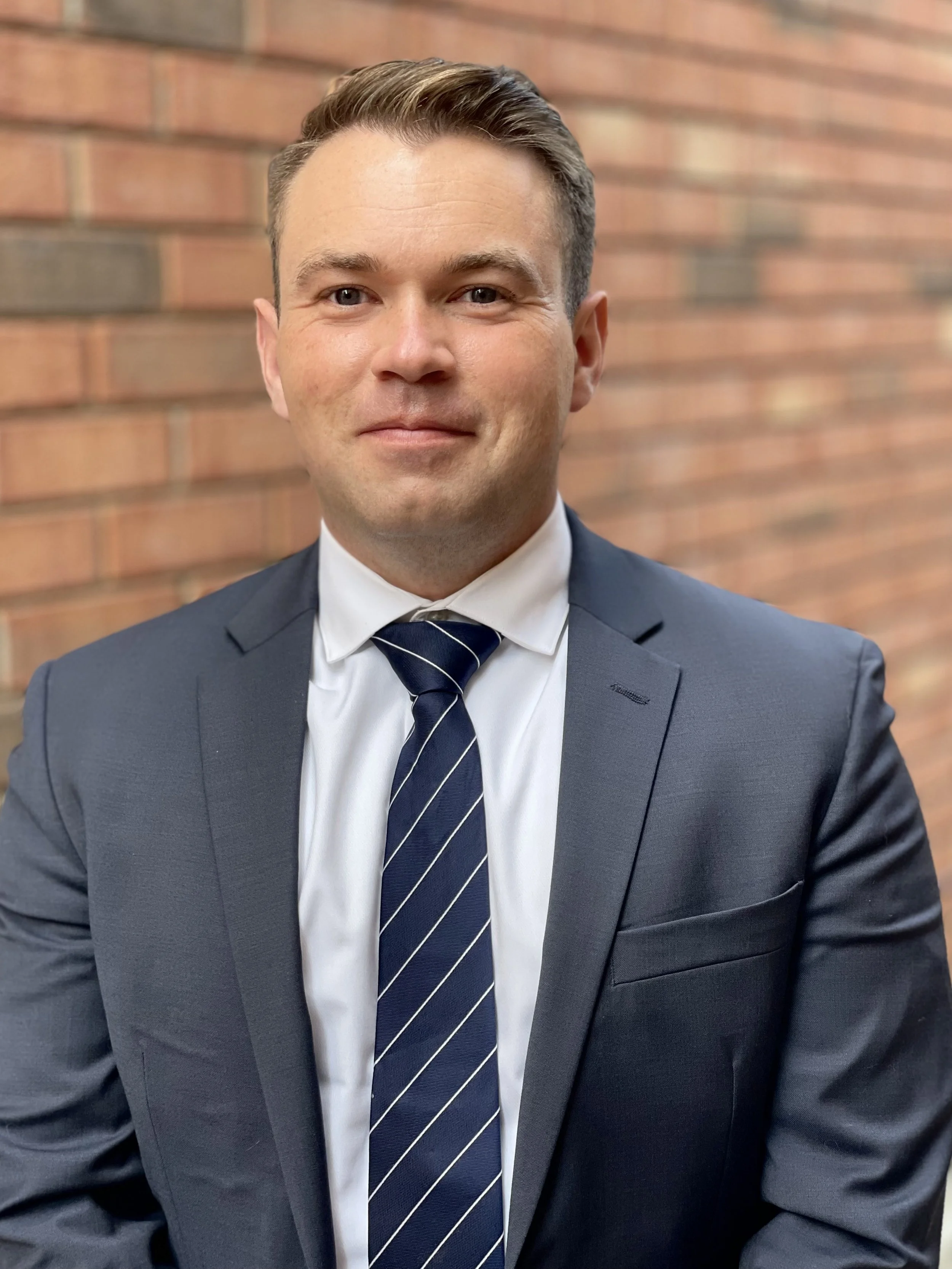 A man in a navy blue suit, white shirt, and striped tie standing in front of a brick wall.