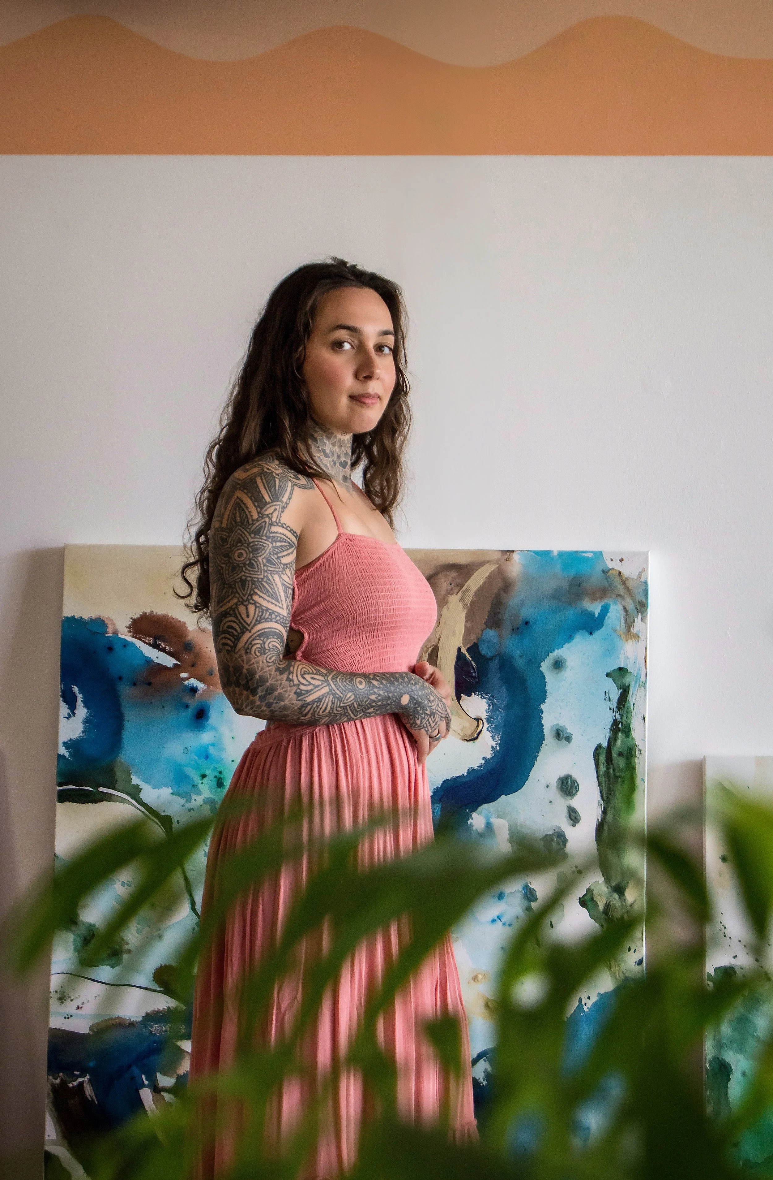 A woman with long wavy hair, tattoos on her left arm and neck, wearing a pink dress, stands indoors in front of an abstract painting with blue, green, and brown colors. Green plants are in the foreground.