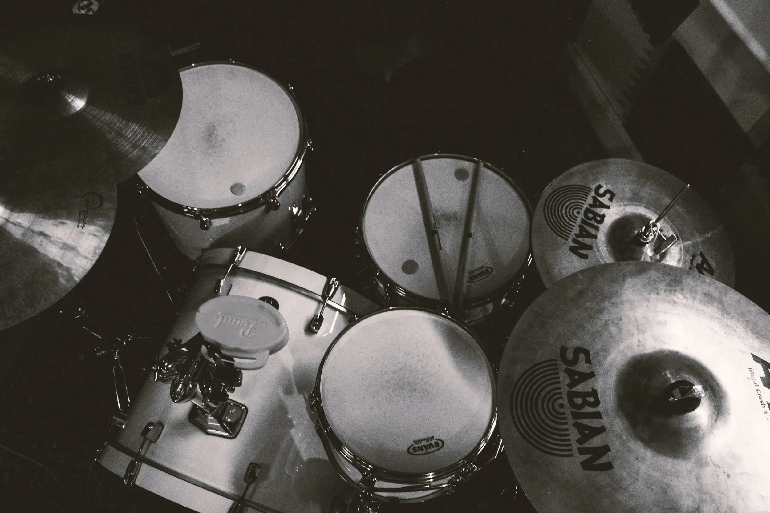 Black and white photo of a drum set with multiple drums and cymbals, some labeled Sabian, with drumsticks resting on one of the drums.