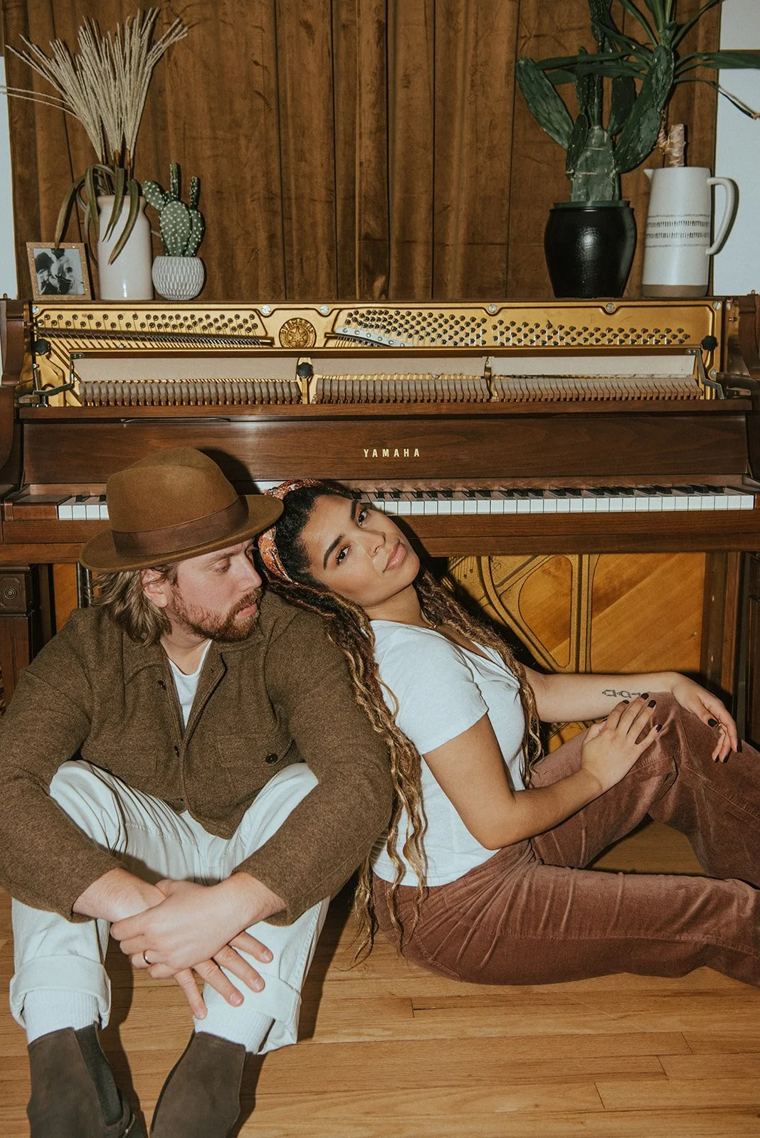 Two people sitting on the floor in front of a vintage upright Yamaha piano, with decorative plants on top, in a room with wood-paneled walls.