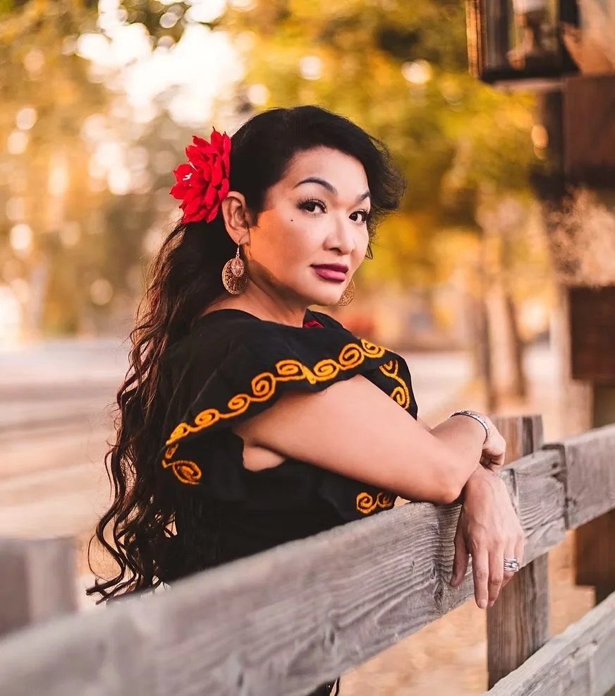 A woman with long dark wavy hair and a red flower in her hair, wearing earrings and a black dress with orange embroidery, leaning on a wooden fence outdoors during autumn.