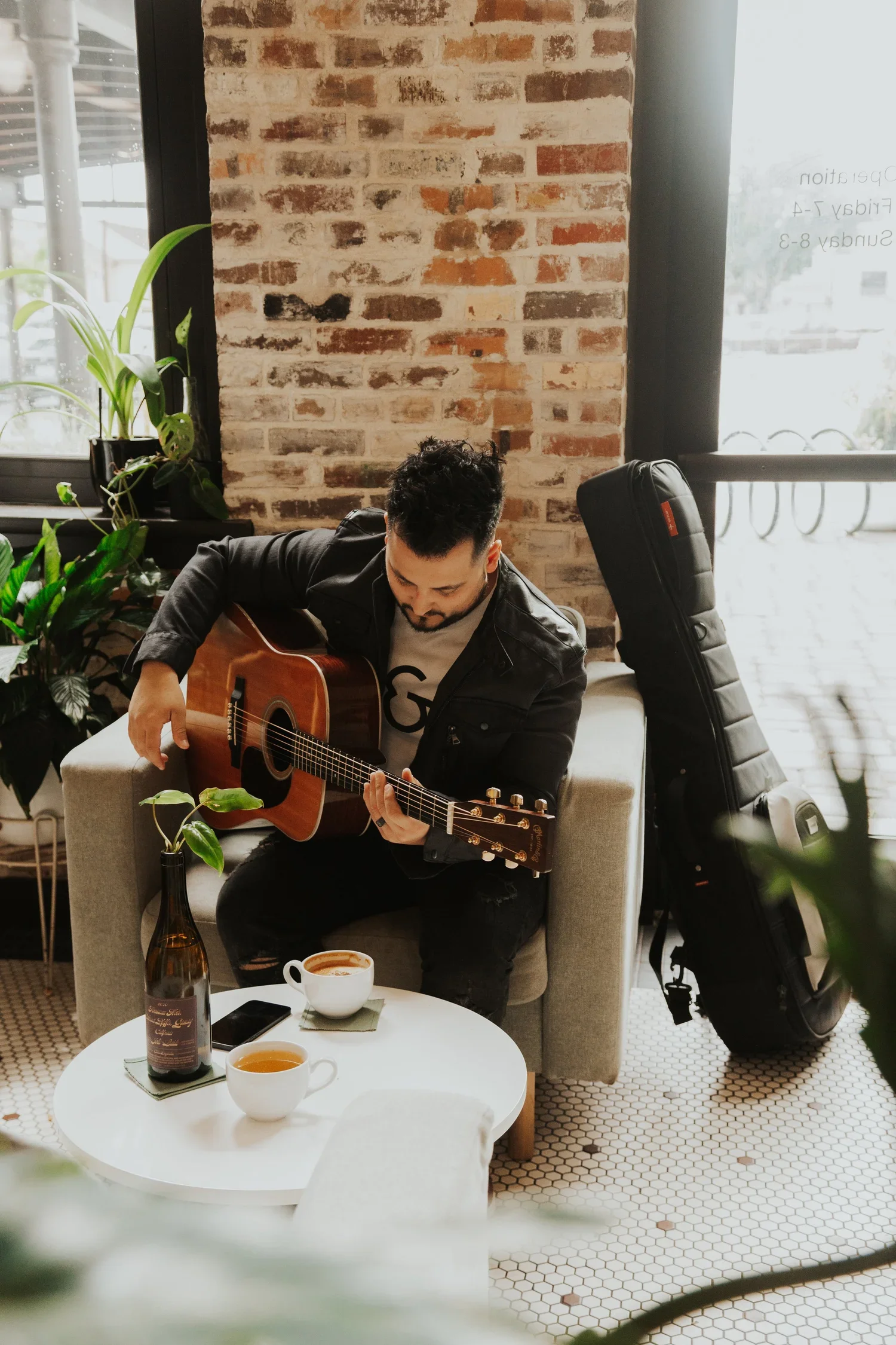 A man playing an acoustic guitar while sitting on a beige armchair in a coffee shop, with cups of coffee on the white round table in front of him, and a guitar case leaning against the wall behind. The setting has a brick wall and large windows with natural light, accompanied by green plants.
