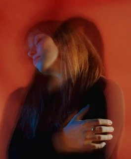 A woman with long brown hair leaning against a red wall, with her eyes closed and arms crossed.