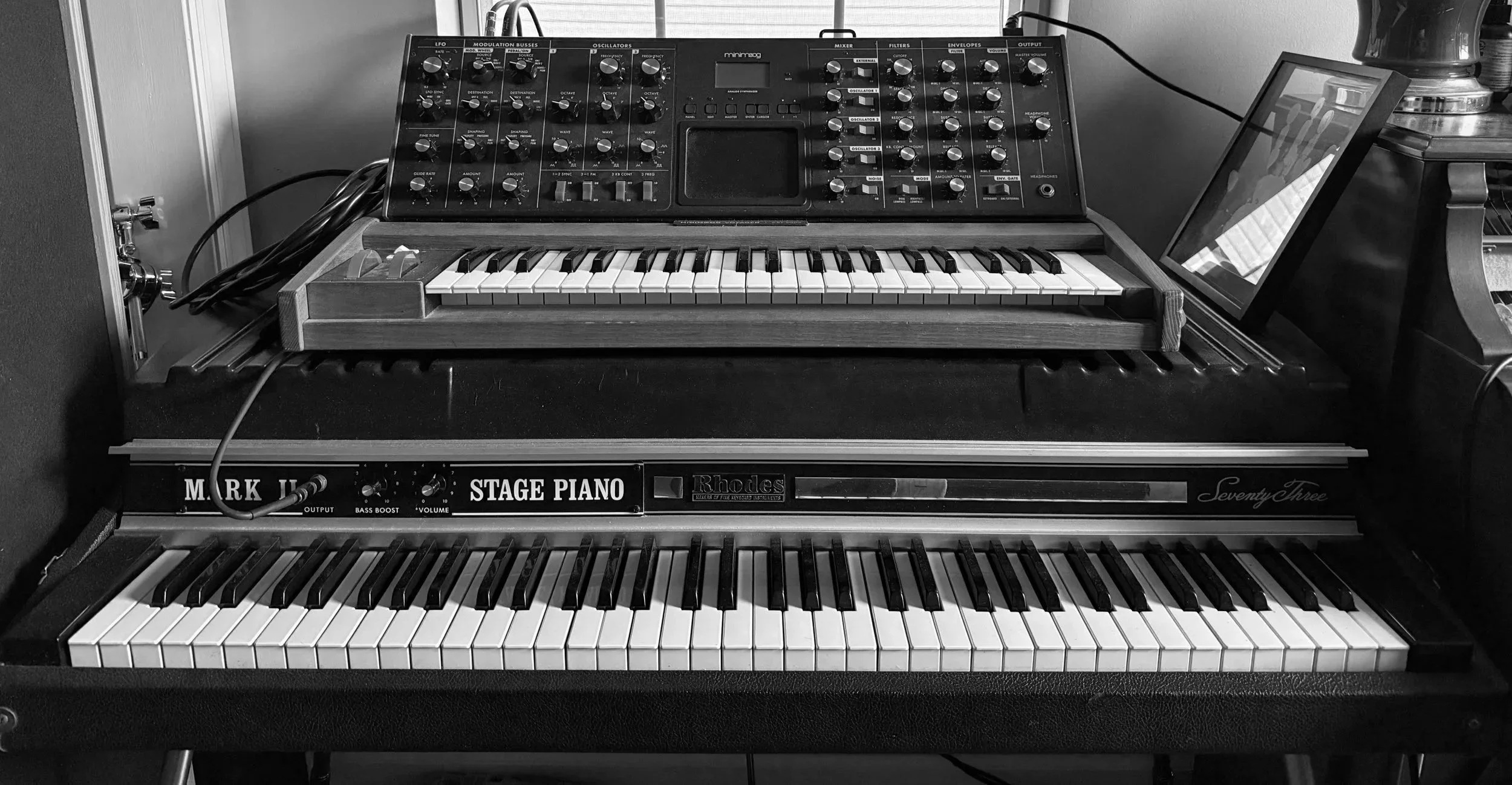 Black and white photograph of two electronic keyboards stacked, with various knobs and controls, on a stand in a music studio or room.