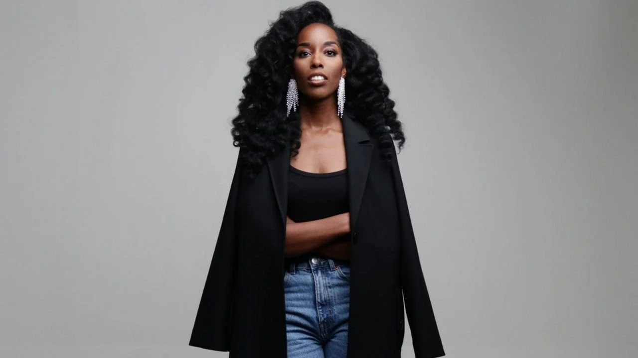 A woman with long, curly black hair wearing a black coat, black top, and blue jeans, standing against a plain gray background.