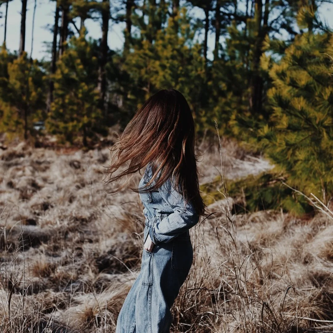 A woman with long brown hair stands in a field of dry grass, facing away with her hands in her pockets, surrounded by green trees.