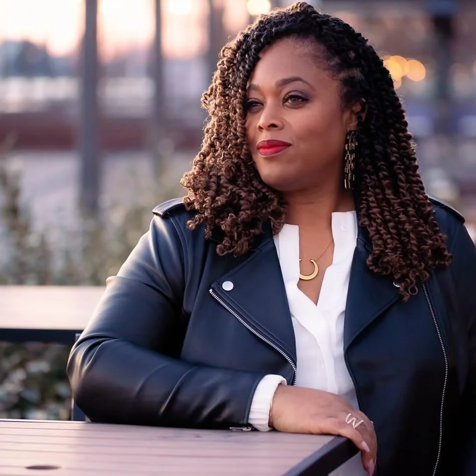 A woman with curly hair and worn red lipstick sitting at a table outdoors during sunset, wearing a white shirt, a black leather jacket, and jewelry including a crescent moon necklace and earrings.
