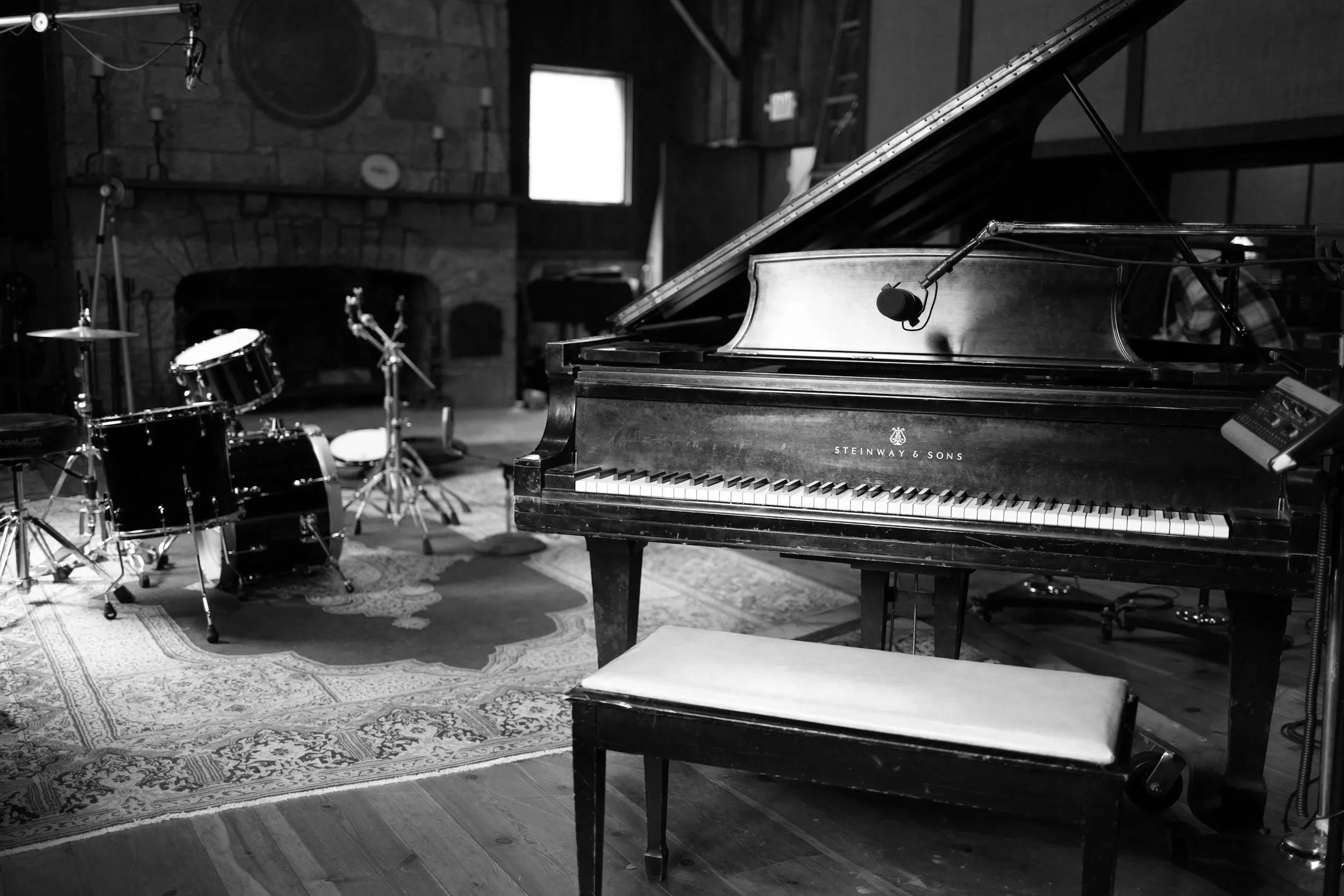 A black and white photo of a music studio with a grand piano in the foreground, and a drum set in the background.