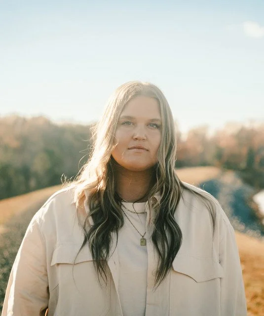 A woman with long blonde hair and a neutral expression standing outdoors in a natural landscape with trees and a dirt path, during daytime.