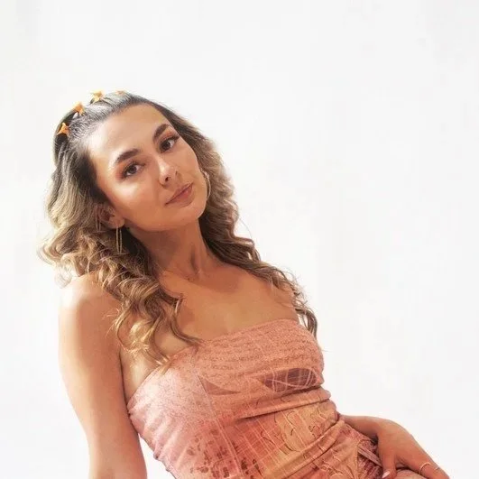 A young woman with wavy hair wearing a strapless pink dress, posing against a white background.