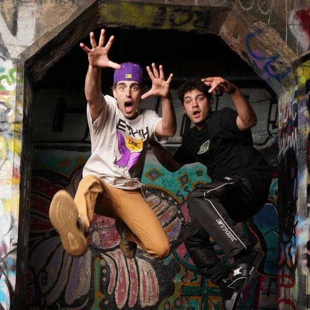 Two young men jumping with surprised expressions in an urban graffiti-covered tunnel.