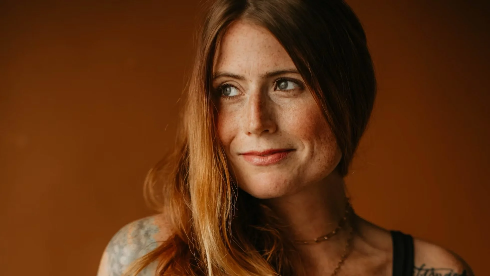 Close-up image of a woman with red hair, freckles, and blue eyes, looking to the left, against a warm brown background.