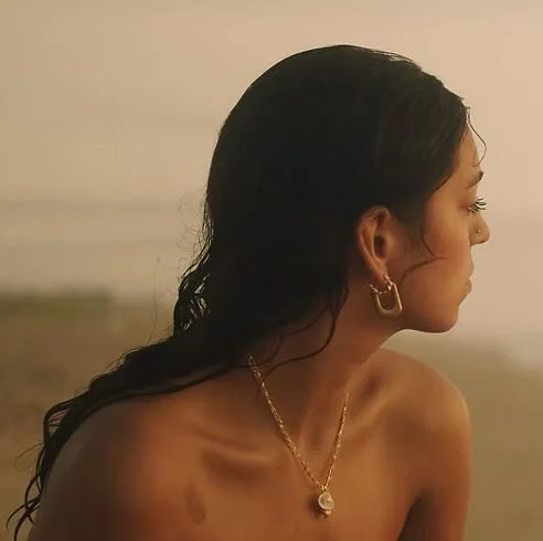 A woman with dark, wavy hair wearing gold hoop earrings and a gold necklace, looking to the right with her bare shoulders visible.