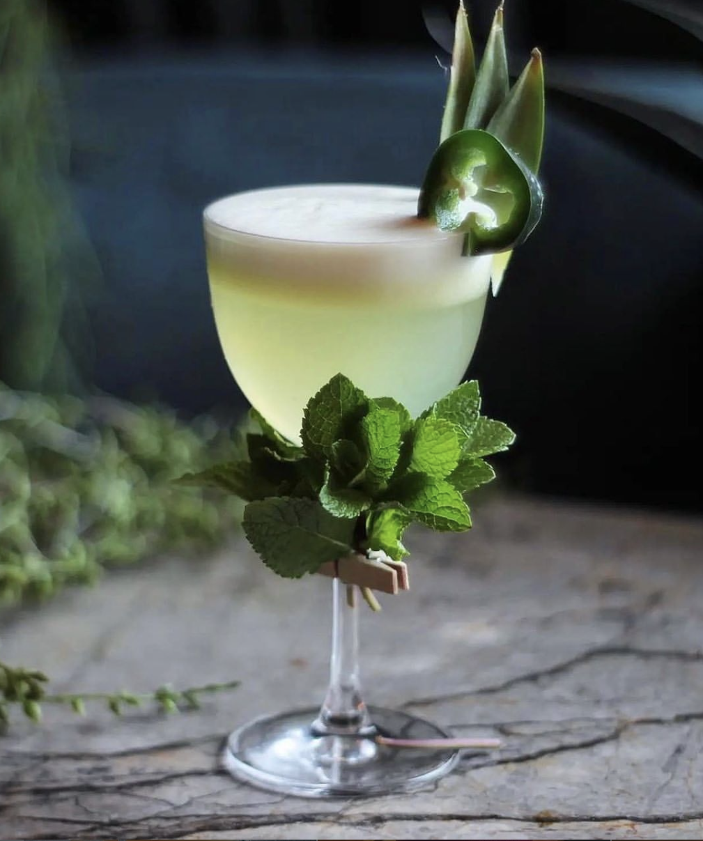 A premium green zero-proof sour by Dry Mixology Arabia, featuring a silky foam, jalapeño slice garnish, and a fresh mint bouquet clipped to the glass stem for a luxury event in the UAE.