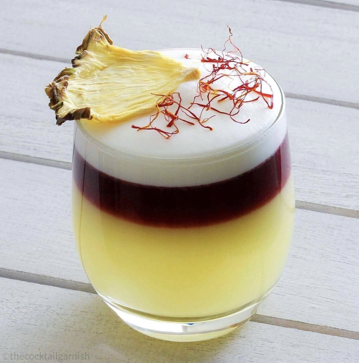 A premium layered zero-proof sour by Dry Mixology Arabia, featuring a crimson red wine float, a frothy egg-white foam, and a garnish of saffron and dehydrated pineapple for luxury events in Dubai.