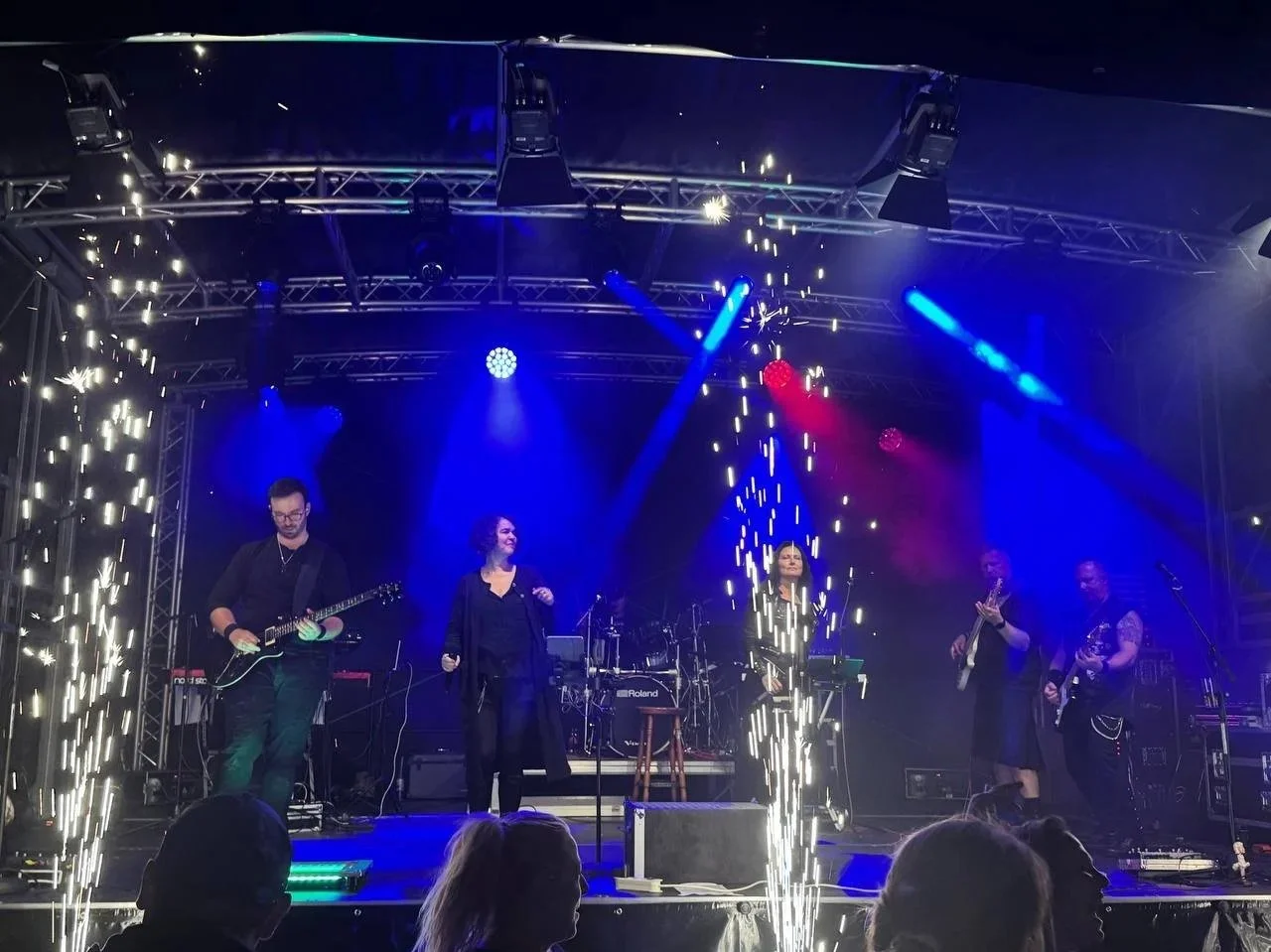 Musikband auf einer Bühne mit blauen und roten Scheinwerfern, Feuerwerk im Vordergrund, Publikum im Vordergrund