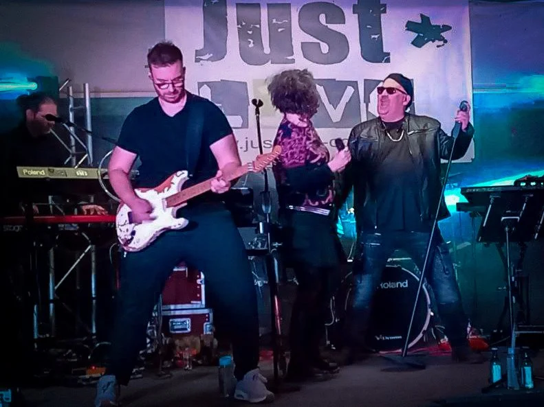 Eine Band tritt auf einer Bühne während eines Live-Auftritts auf, mit einem Hintergrundschild mit dem Wort "Just Live". Es gibt drei Musiker, einer mit E-Gitarre, einer singend und einer mit einem Mikrofon. Die Beleuchtung ist bunt.