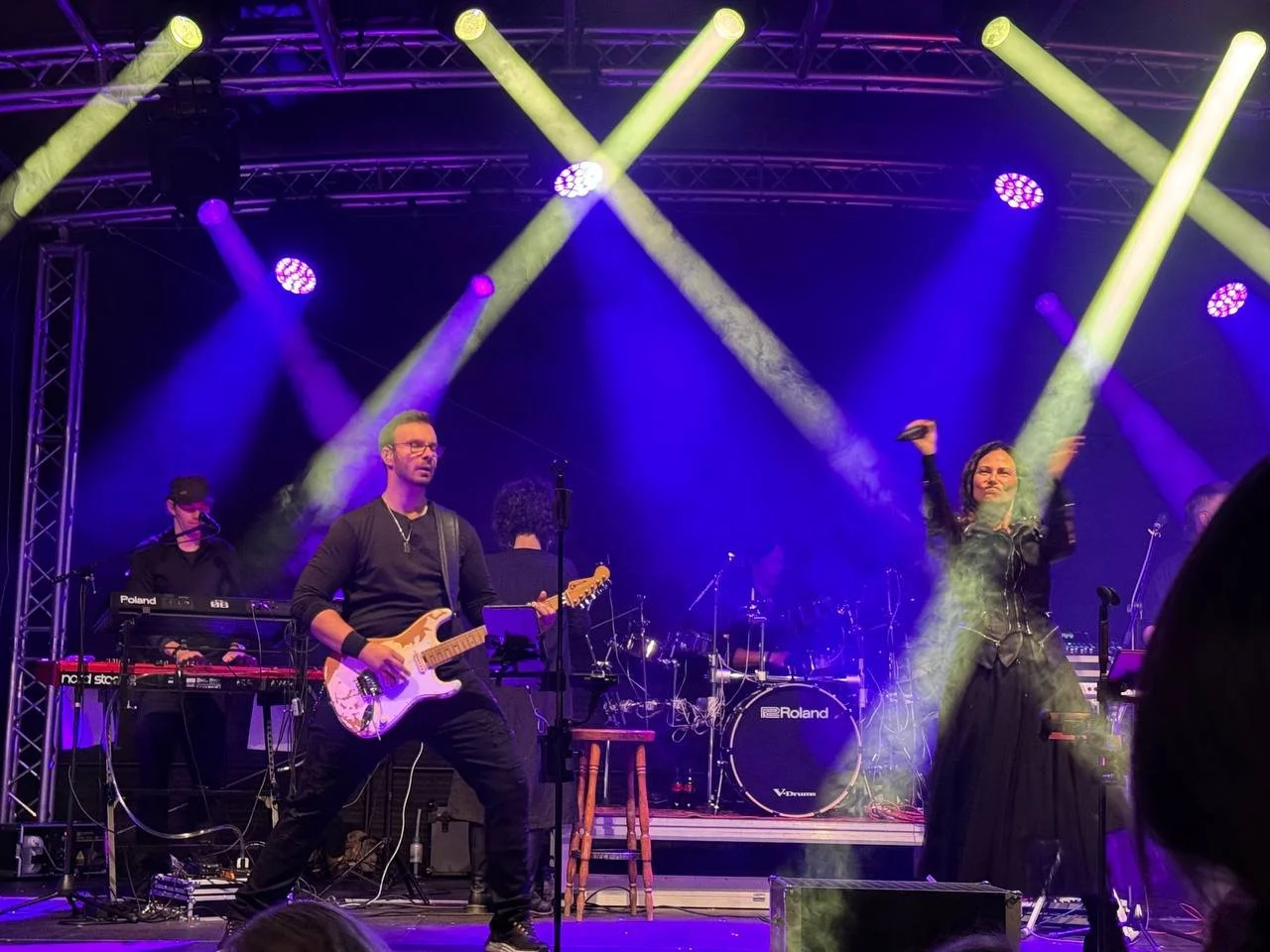 Musiker auf einer Bühne bei einem Konzert, umgeben von Lichtstrahlen in Gelb und Lila, mit Gitarren, Tastaturen und Schlagzeug.