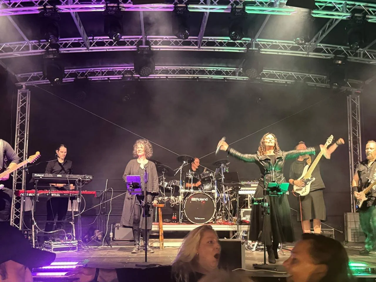 Musiker auf einer Bühne während eines Konzerts, die Band besteht aus mehreren Mitgliedern, einige spielen Gitarre, ein Schlagzeug ist im Hintergrund, bunte Bühnenbeleuchtung, das Publikum ist im Vordergrund sichtbar.