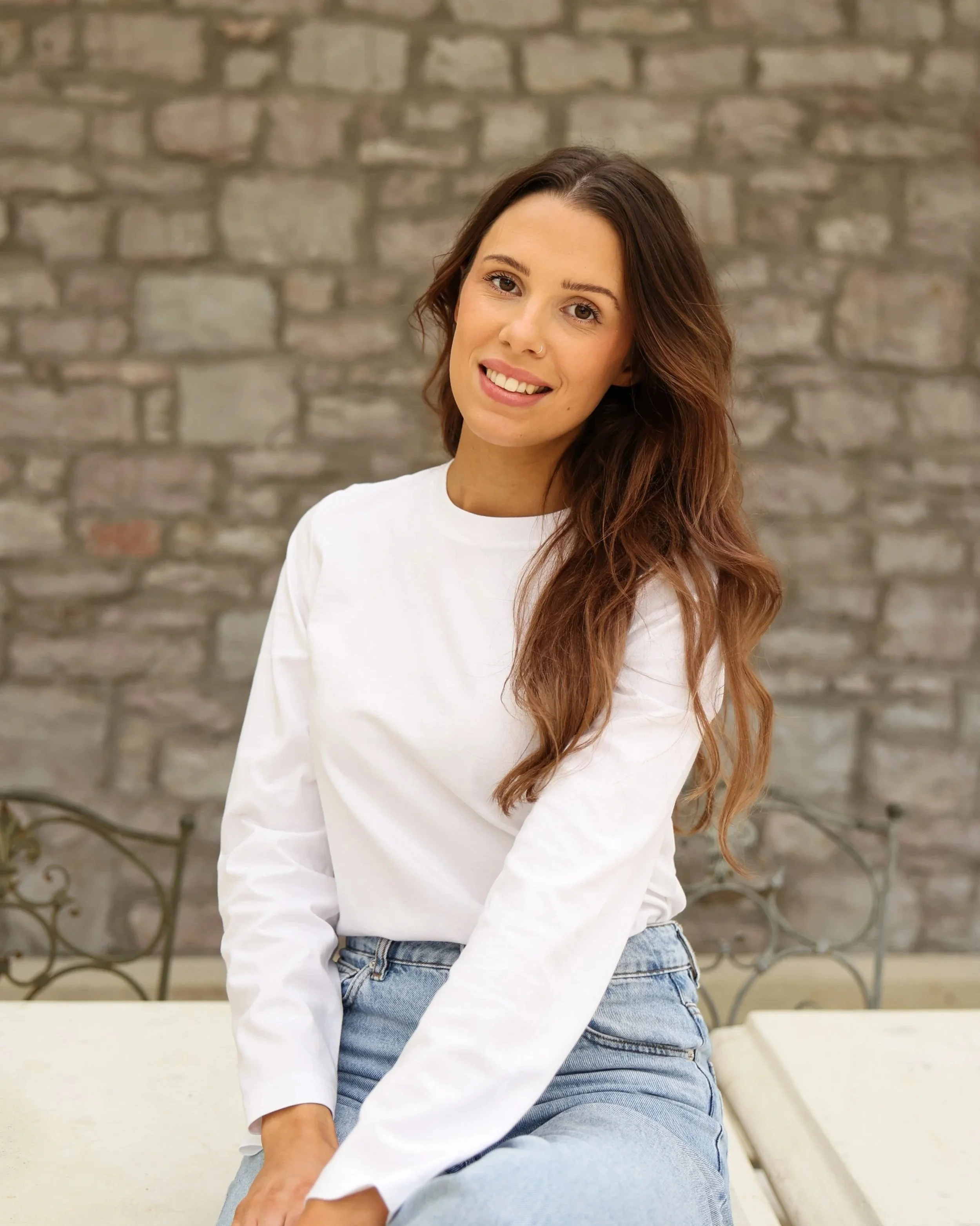A woman with brown wavy hair and a nose piercing smiling, wearing a white long-sleeve shirt and light blue jeans, sitting outdoors in front of a brick wall with a black wrought iron bench in the background.