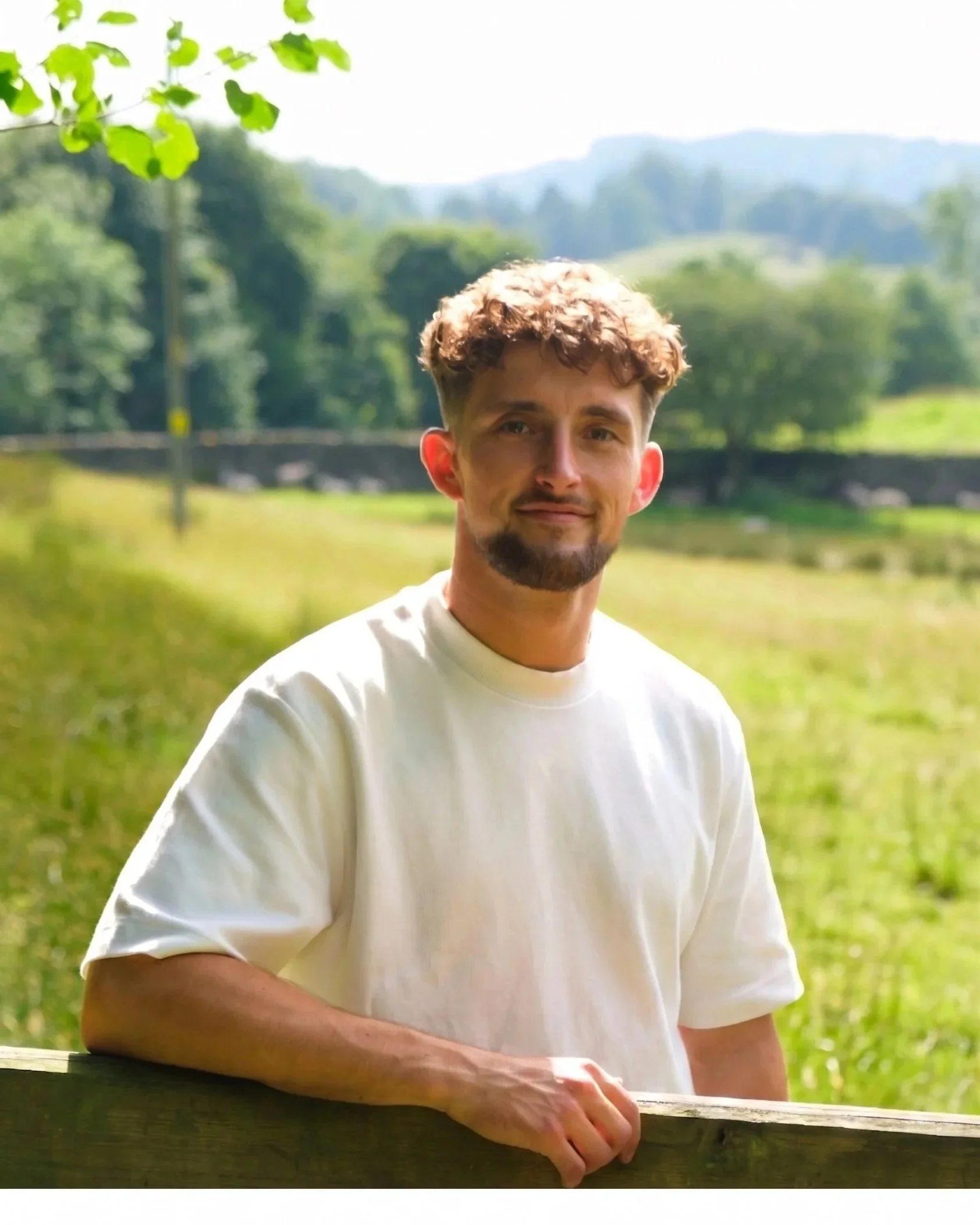 A portrait of a young man with short, curly brown hair and a beard, smiling slightly, standing outdoors in a green, natural setting with trees and hills in the background, wearing a white t-shirt.