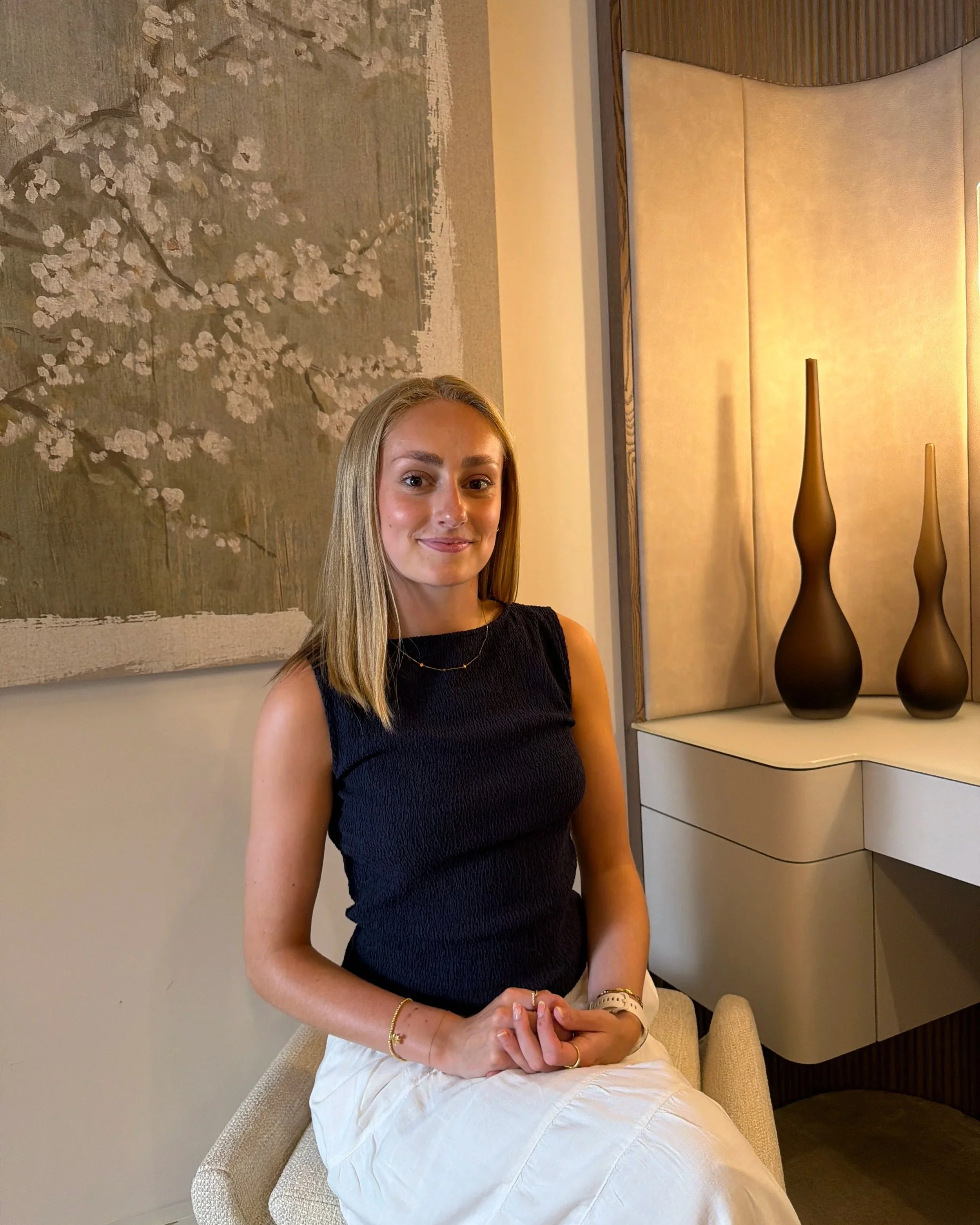 A woman sitting on a beige chair in front of a wall with a large floral artwork, wearing a black sleeveless top, with vases and decorative objects on a white cabinet beside her.