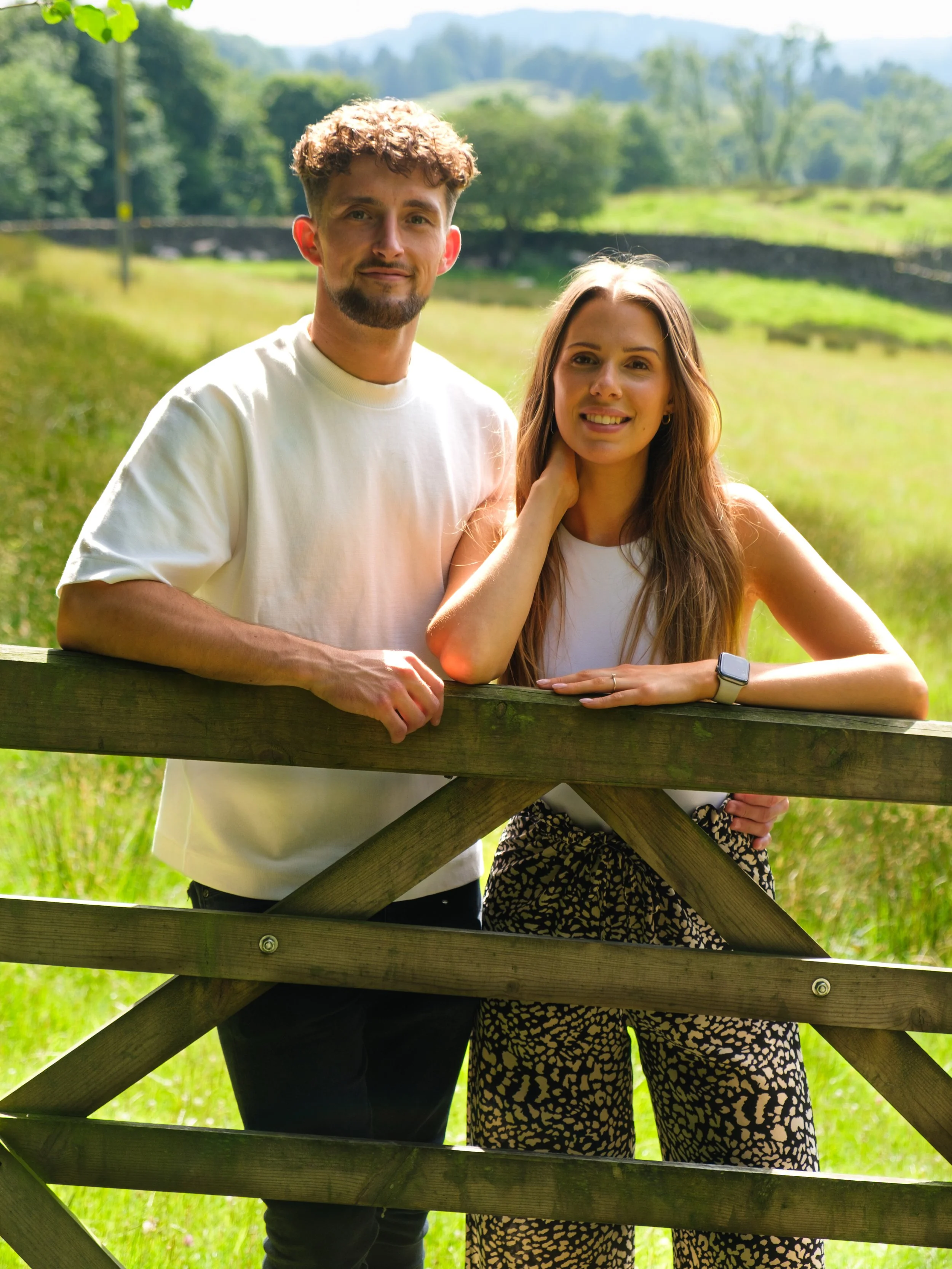 A young man and woman standing outdoors by a wooden fence, smiling at the camera with a lush green landscape and trees in the background.