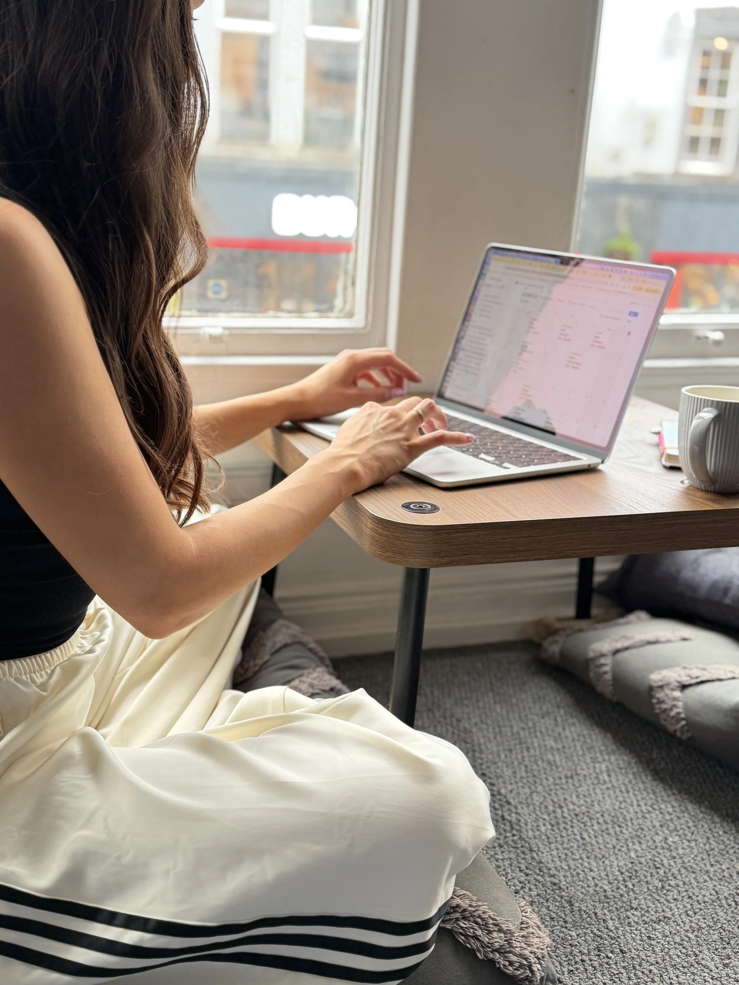 Person sitting by a window using a laptop, with a mug and phone on the table, and slippers on a rug nearby.