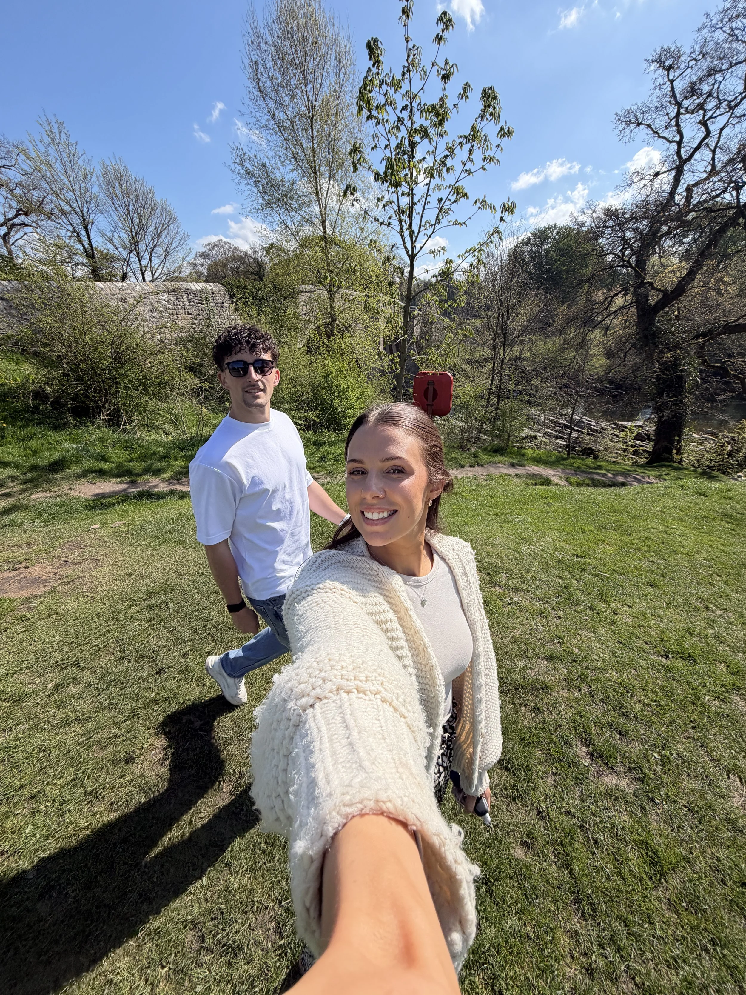 A woman and a man standing outdoors in a park on a sunny day with blue skies and trees in the background. The woman is taking a selfie, and the man is standing behind her, smiling and wearing sunglasses.