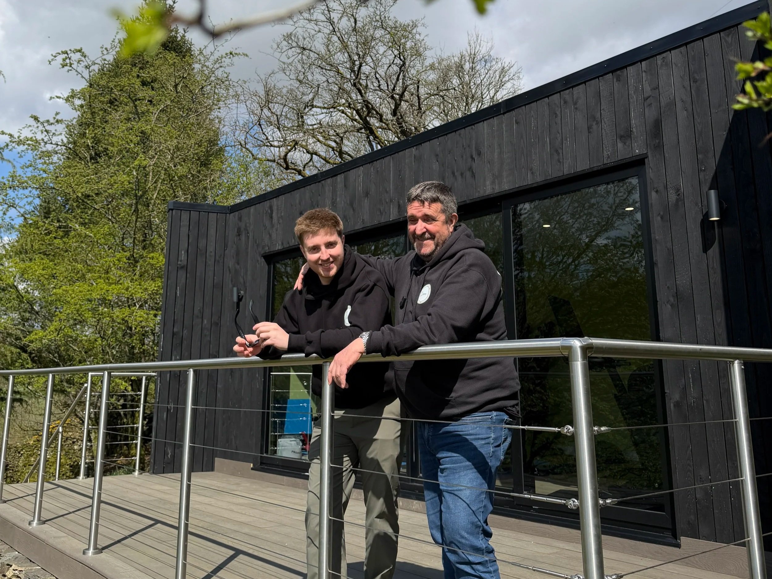 Two men standing on a wooden deck outside a modern black house, smiling and posing with their arms around each other. Green trees and a partly cloudy sky are in the background.