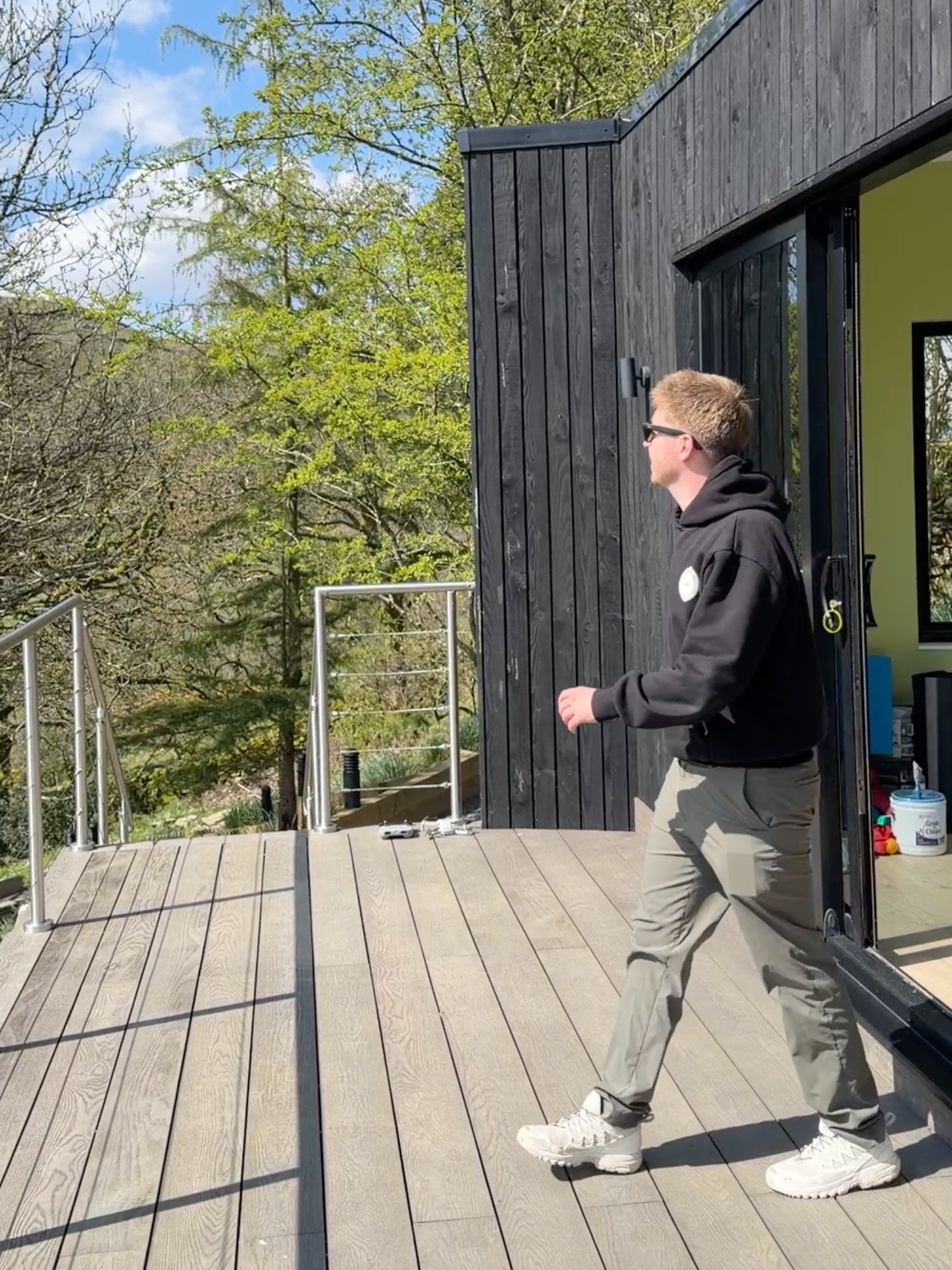 A young man wearing sunglasses, a black hoodie, beige pants, and white sneakers walking on a wooden deck outside a modern building with black wooden siding and a bright yellow interior wall. There are trees with green leaves and a partly cloudy sky in the background.