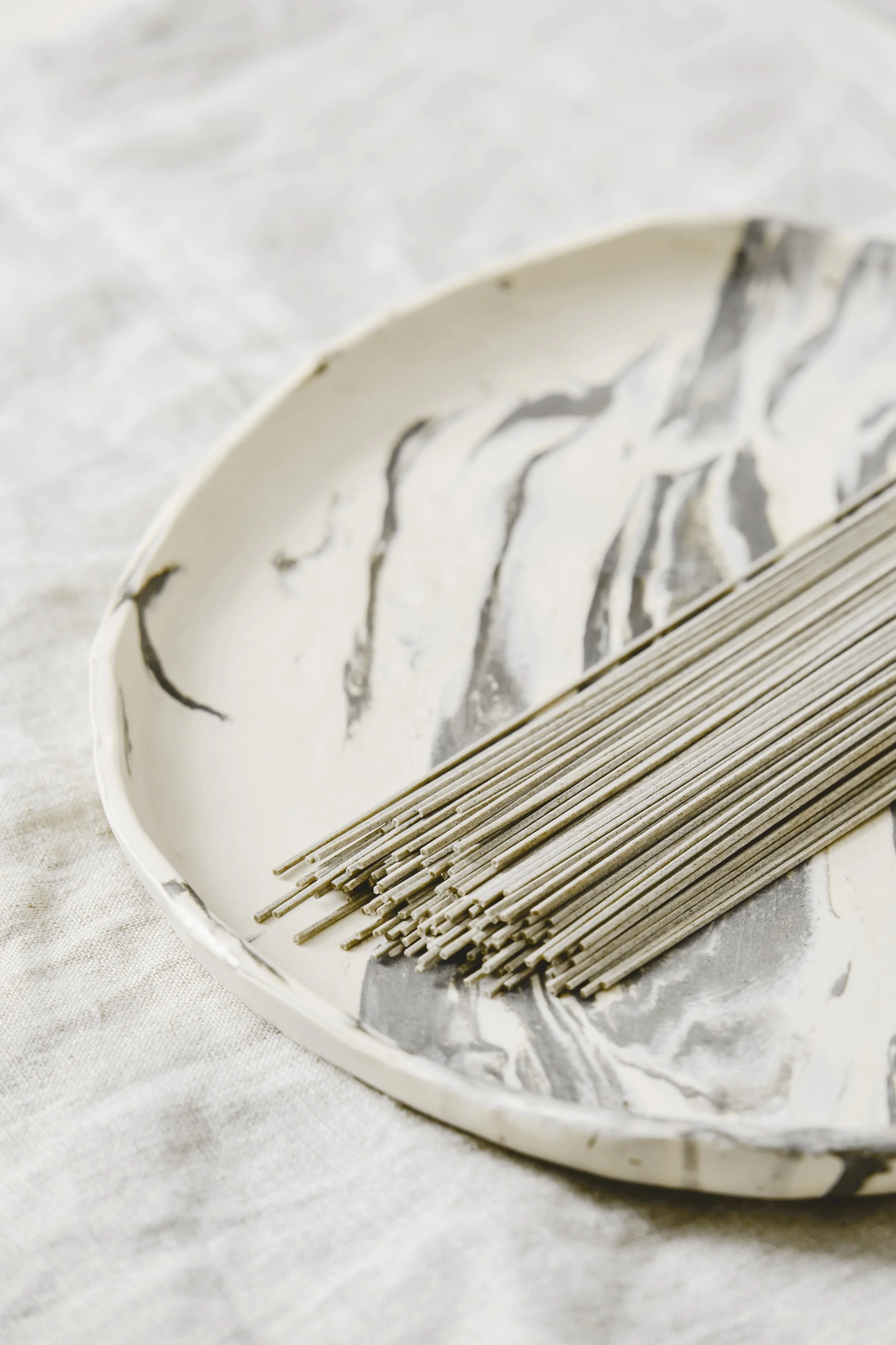 Uncooked soba noodles on a ceramic plate with black streak pattern, placed on a light-colored fabric surface.