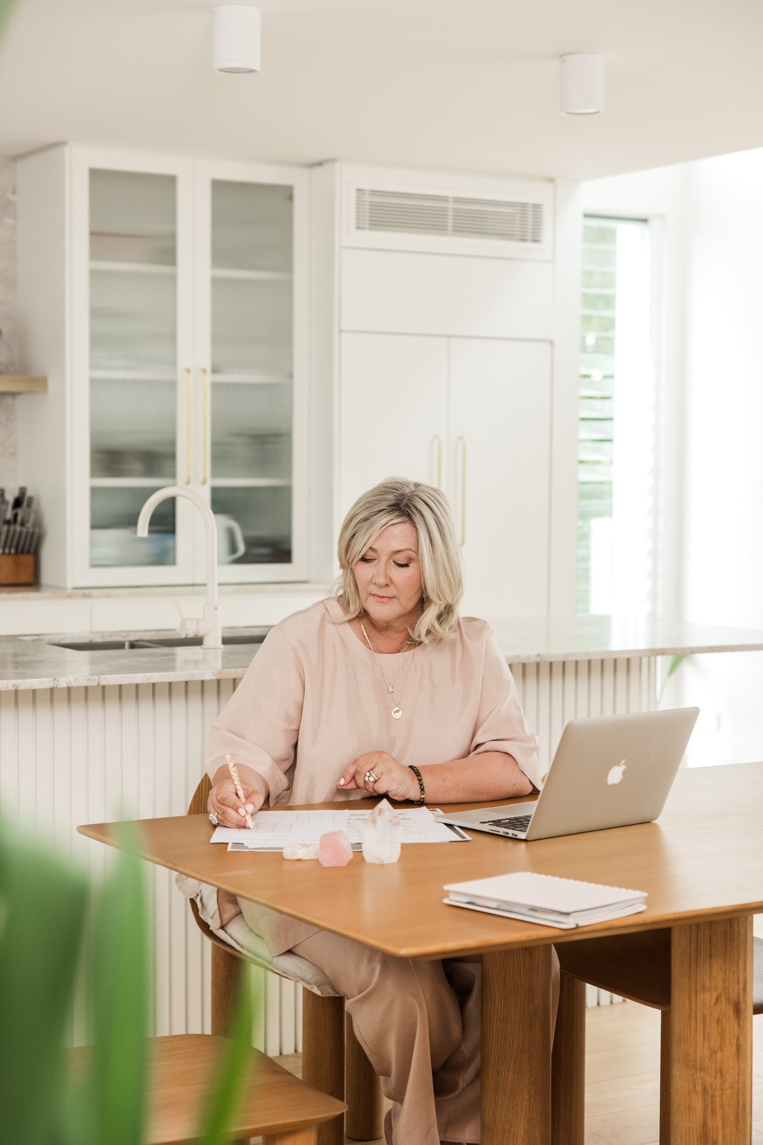 A woman sitting at a wooden table working on papers with a laptop open in front of her in a bright kitchen.