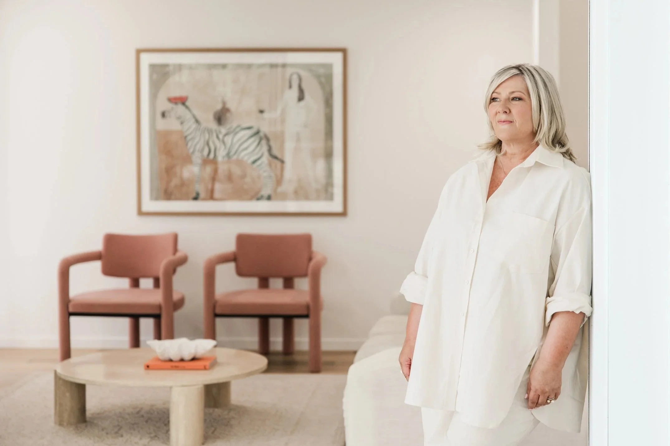 A woman with gray hair wearing a white shirt stands in a modern, minimalistic living room with pastel pink chairs, a light-colored round coffee table, and artwork featuring a zebra on the wall.