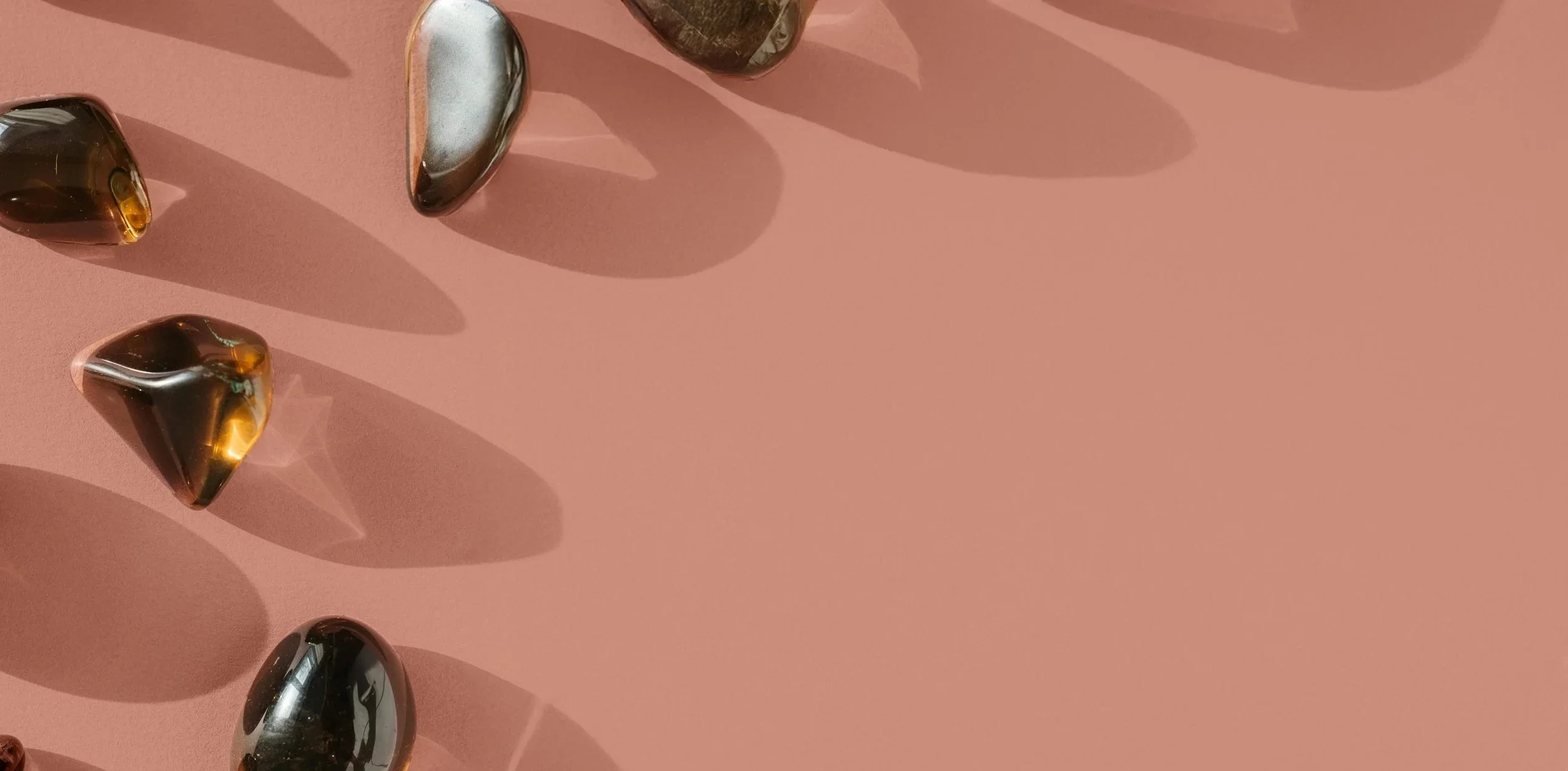 Multiple polished stones of various shapes and colors, including brown, gray, and black, arranged on a pink surface, casting elongated shadows.